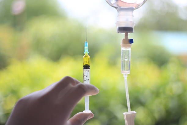 Hand holding syringe with yellow liquid, next to IV drip bag, outdoor setting.