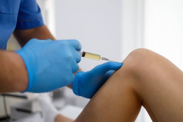 Person in blue gloves injecting a knee with a syringe, likely for a medical procedure in a clinic.