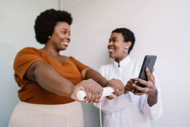 Two Black women smiling, one exercising with a device while the other holds a phone.