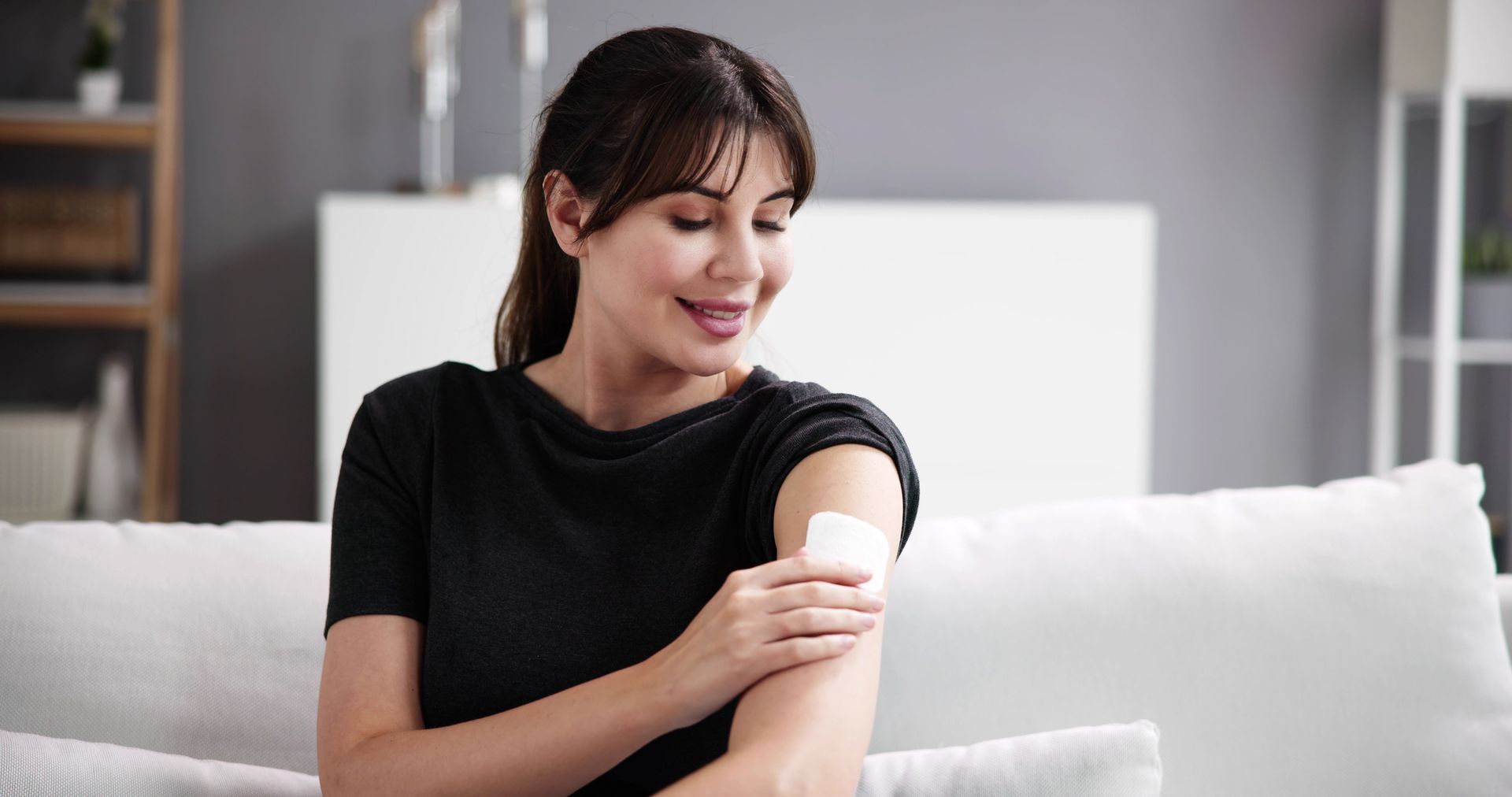 Woman with dark hair looking at medical patch on arm, smiling, indoors.
