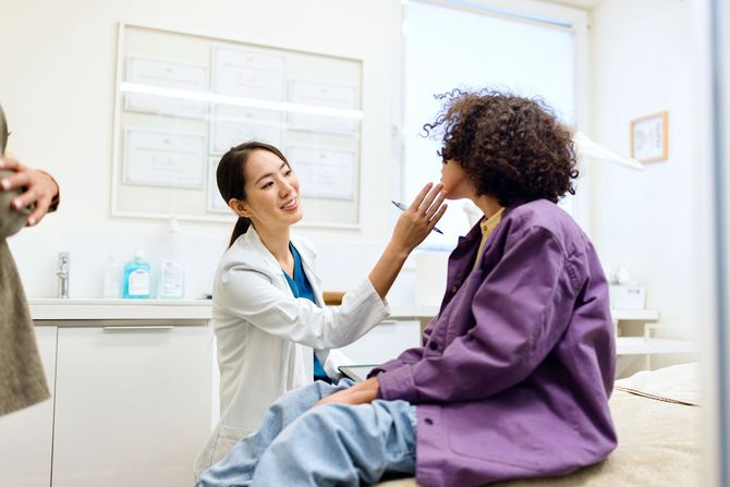Doctor examines patient's throat in an exam room. The doctor is Asian and the patient is Black.