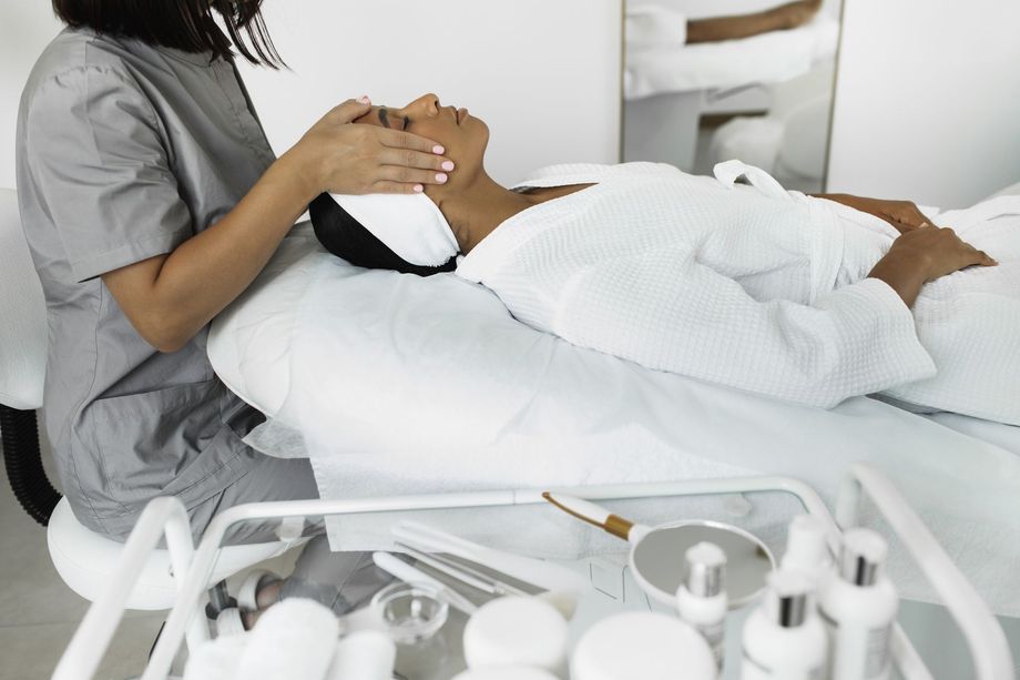 Person receiving facial treatment at a spa, face-down on a bed. Practitioner in scrubs. White robe and towel, various skincare products.
