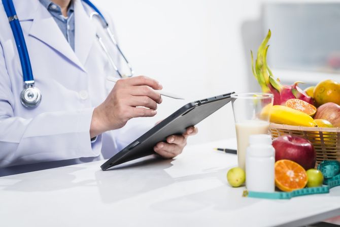 Doctor in lab coat, using tablet, with fruits, smoothie, and supplements on desk.