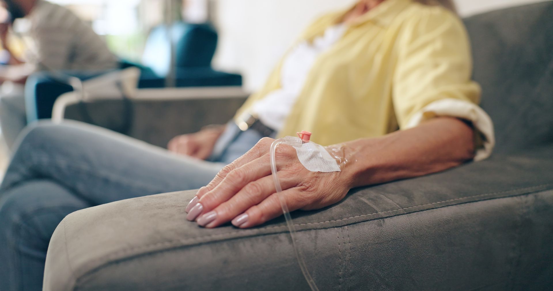 Woman receiving IV infusion, seated in chair. Yellow shirt, blue jeans, light skin, indoor setting.