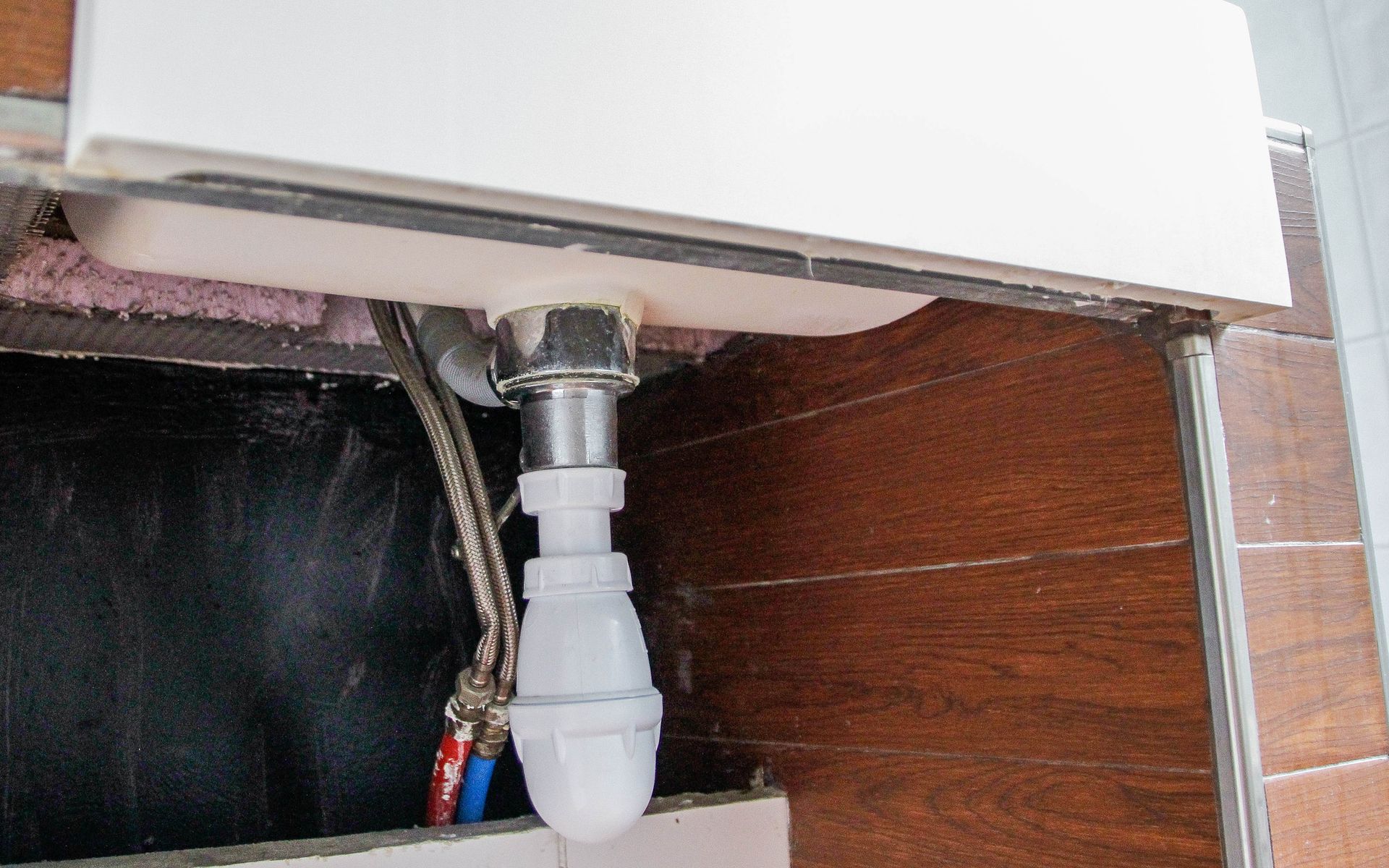A close-up view of a kitchen sink drainpipe with plumbing connections and wooden paneling.