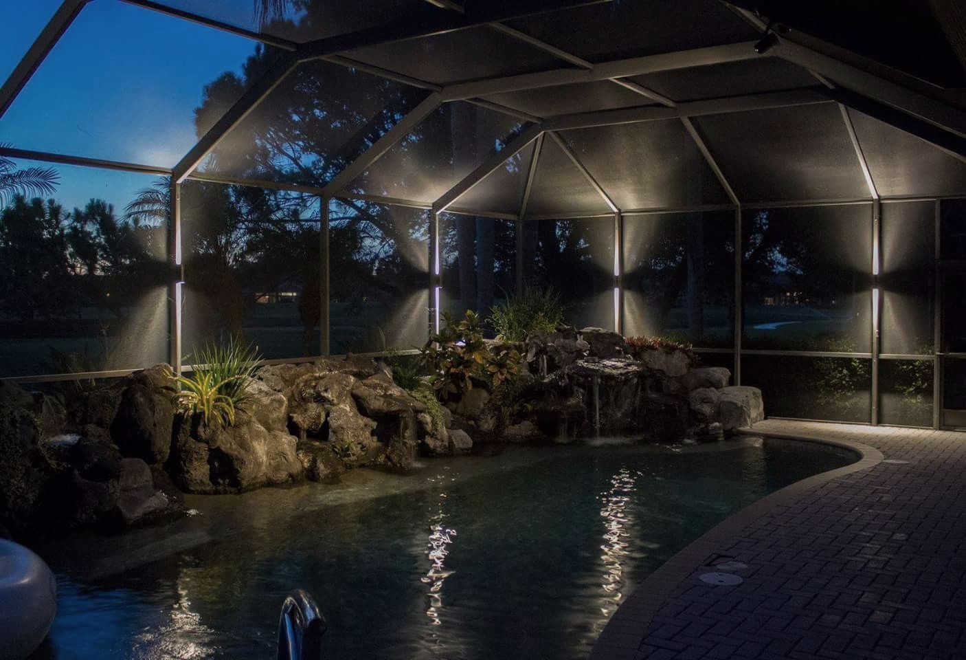 A swimming pool is lit up at night in a screened in area.