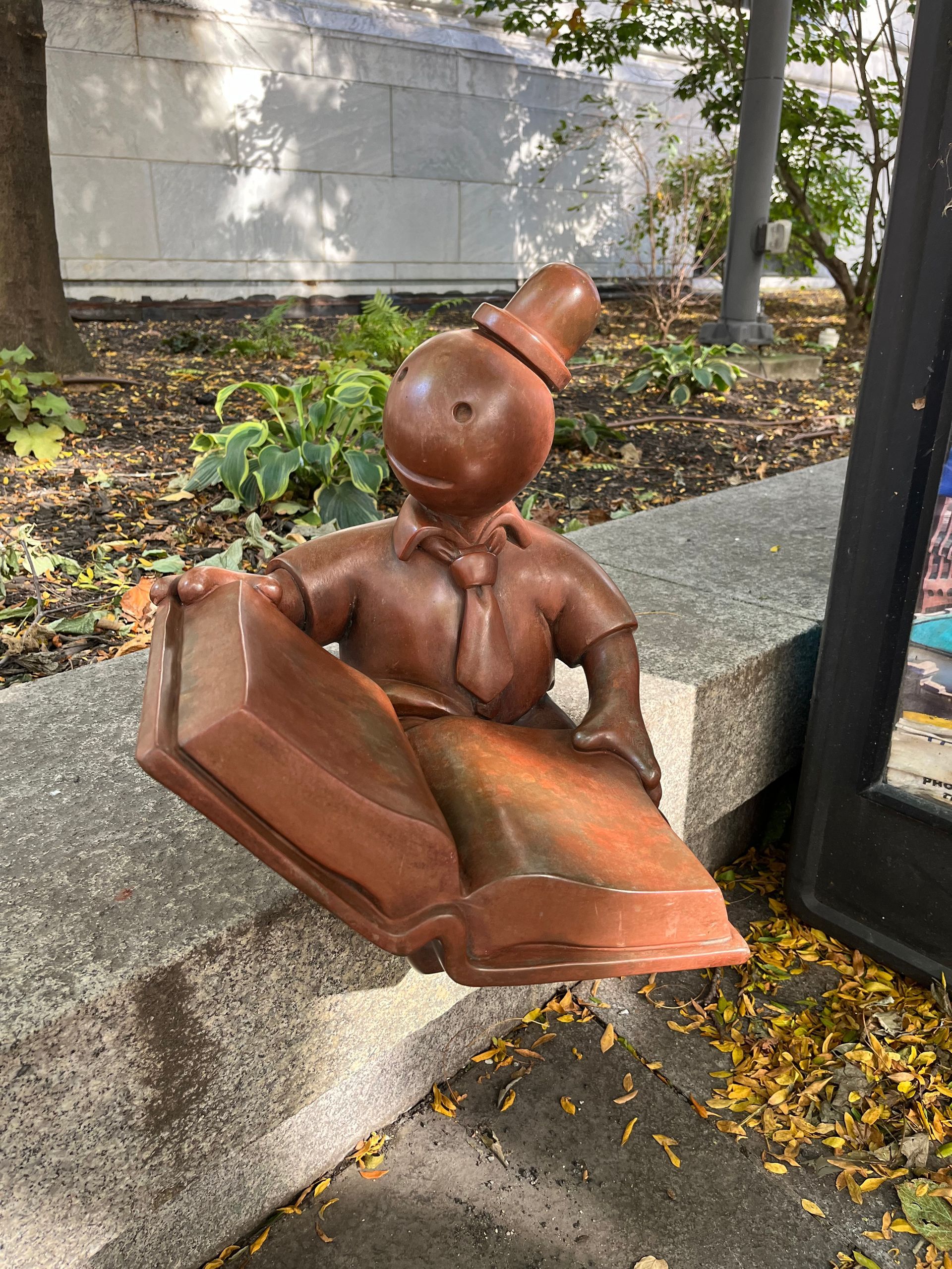 A brown, stylized sculpture of a figure wearing a top hat and tie while reading a large, open book on a stone ledge.