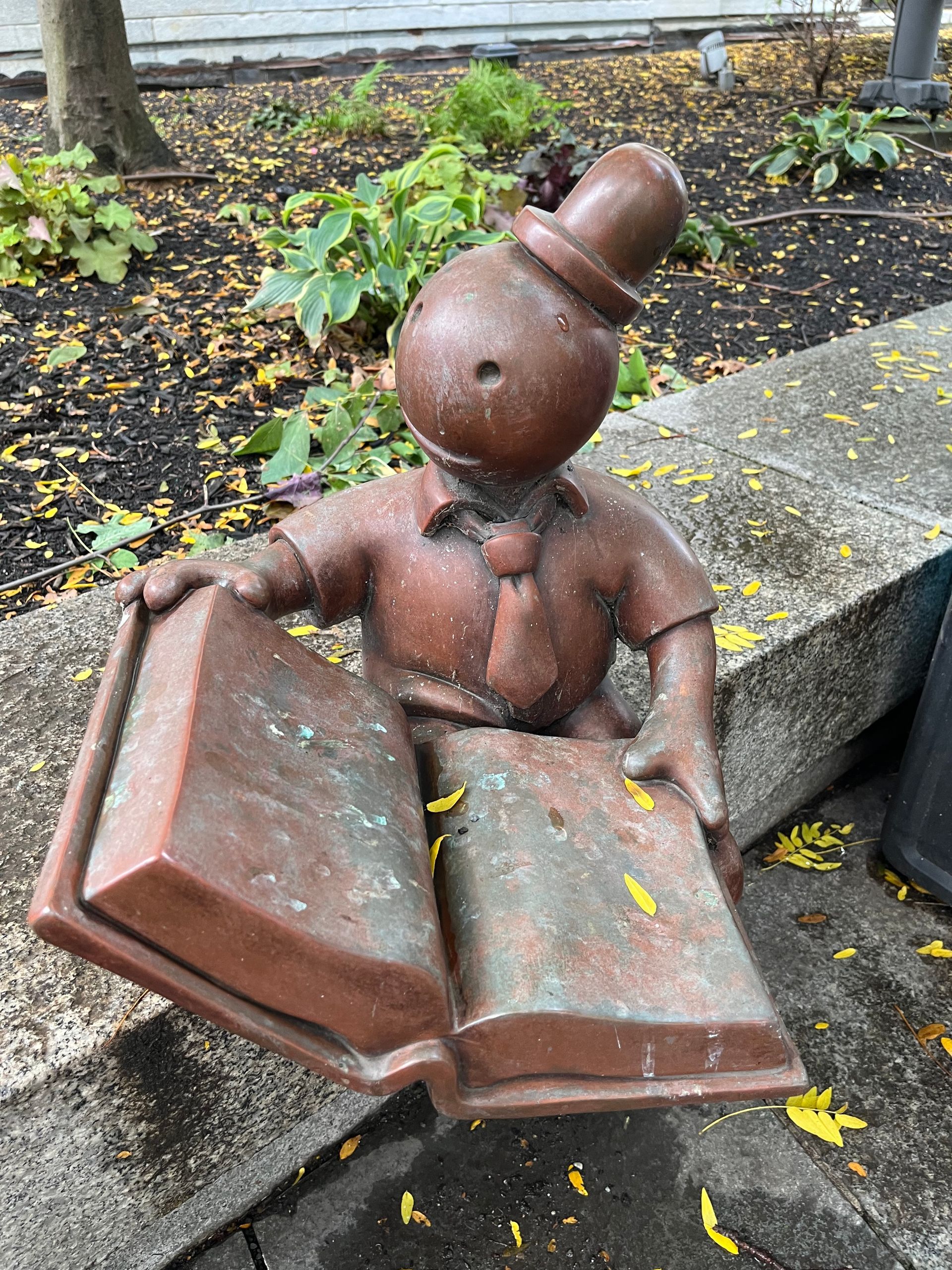 A brown, spherical-headed statue wearing a top hat and tie, sitting and reading an open book on a stone ledge.