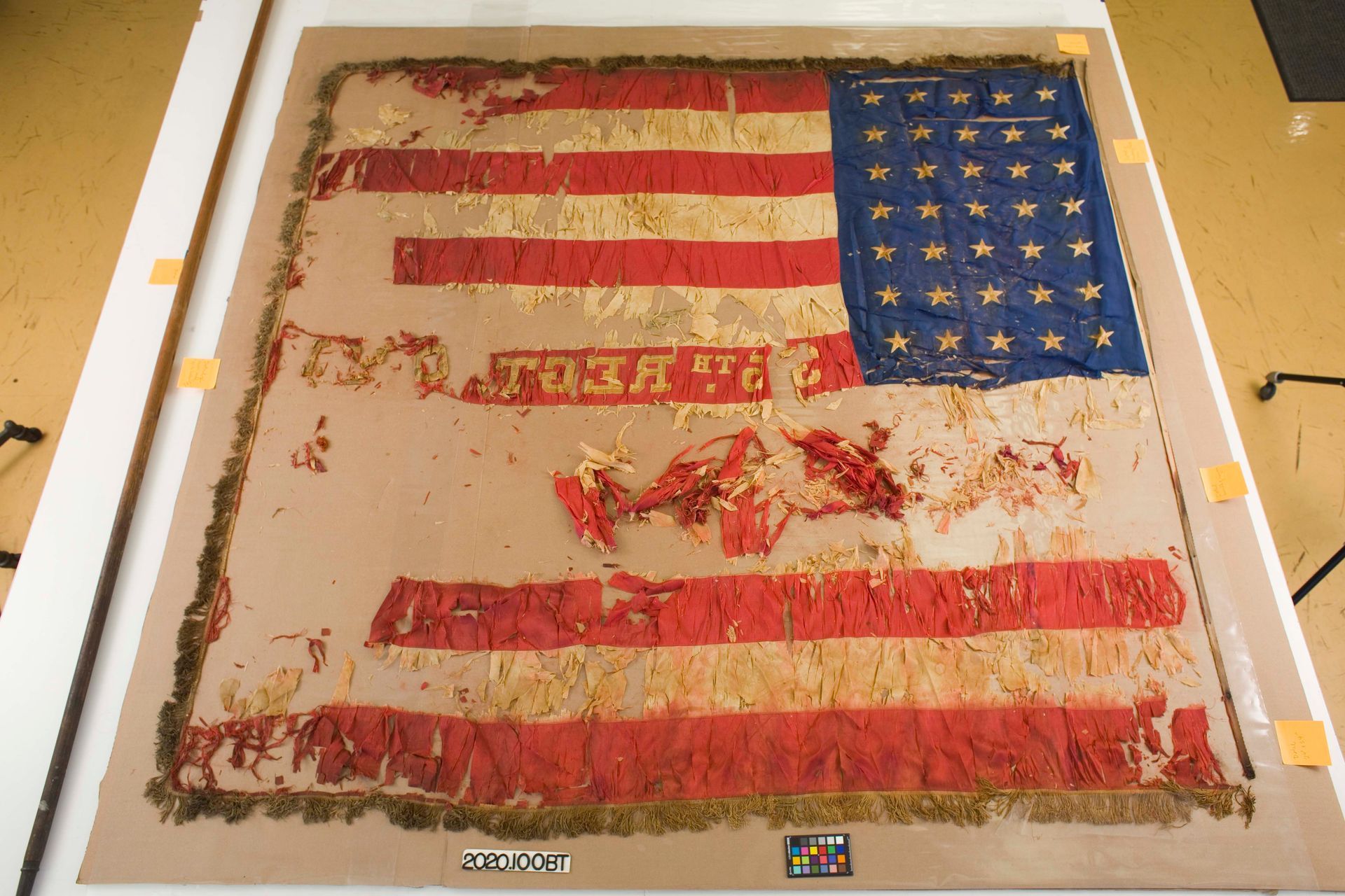 An aged, fragmented US flag with visible stitching and worn fabric is mounted flat on a light brown display board.