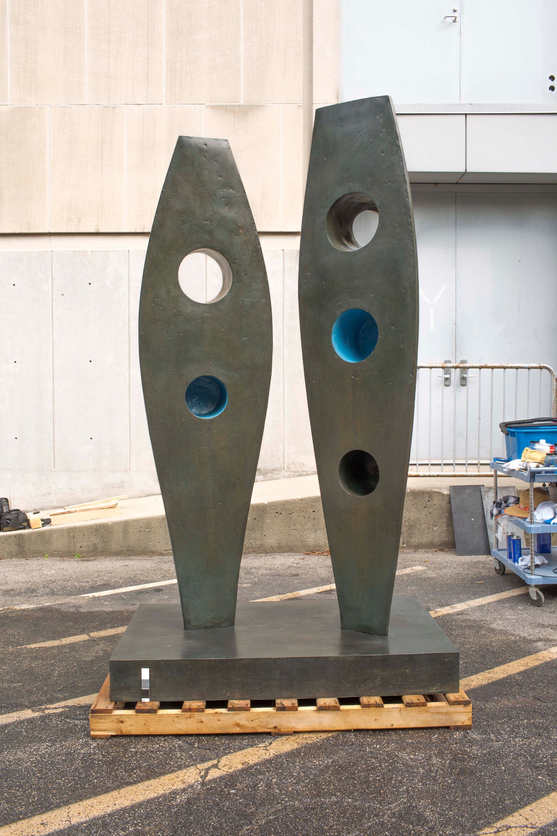 A dark, two-piece abstract metal sculpture with circular cutouts featuring blue accents, set on a base outdoors.