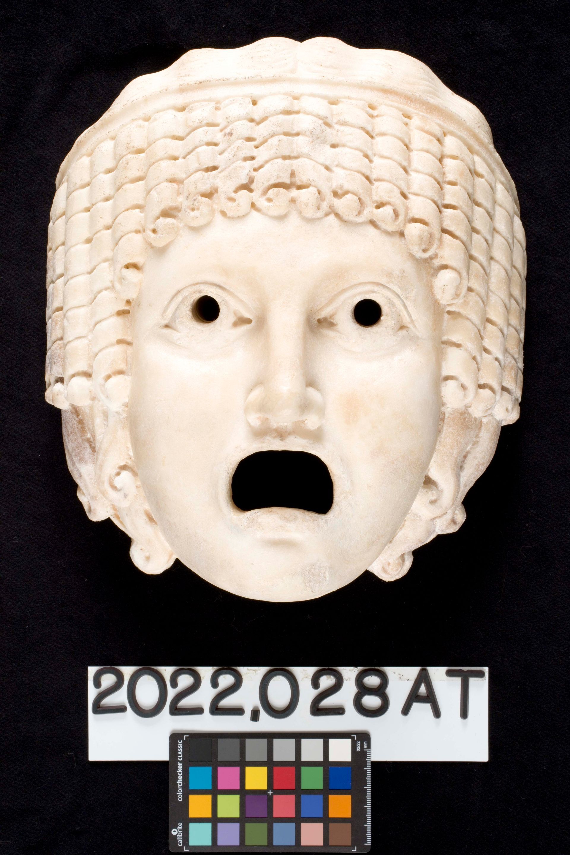 A white marble mask of a classical theatrical character with ringlet curls, wide open eyes, and an open, gaping mouth.