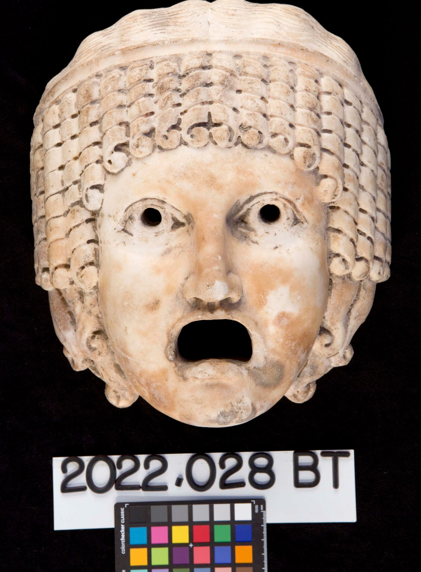 A pale, stone theatrical mask with curled hair and an open, gaping mouth, against a black background.