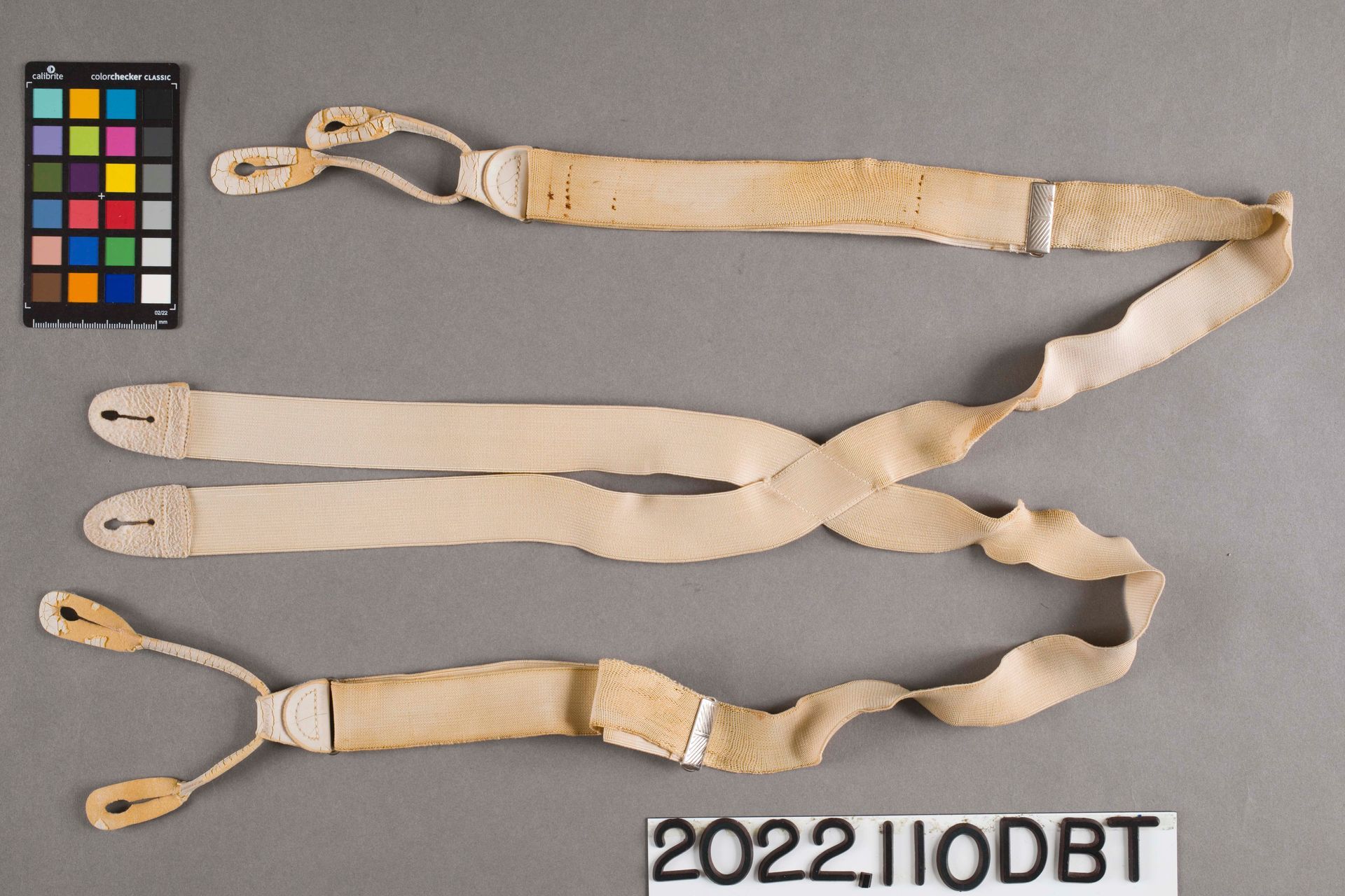 Off-white suspenders with button-tab ends and a metal adjuster, laid flat against a gray background with a color card.