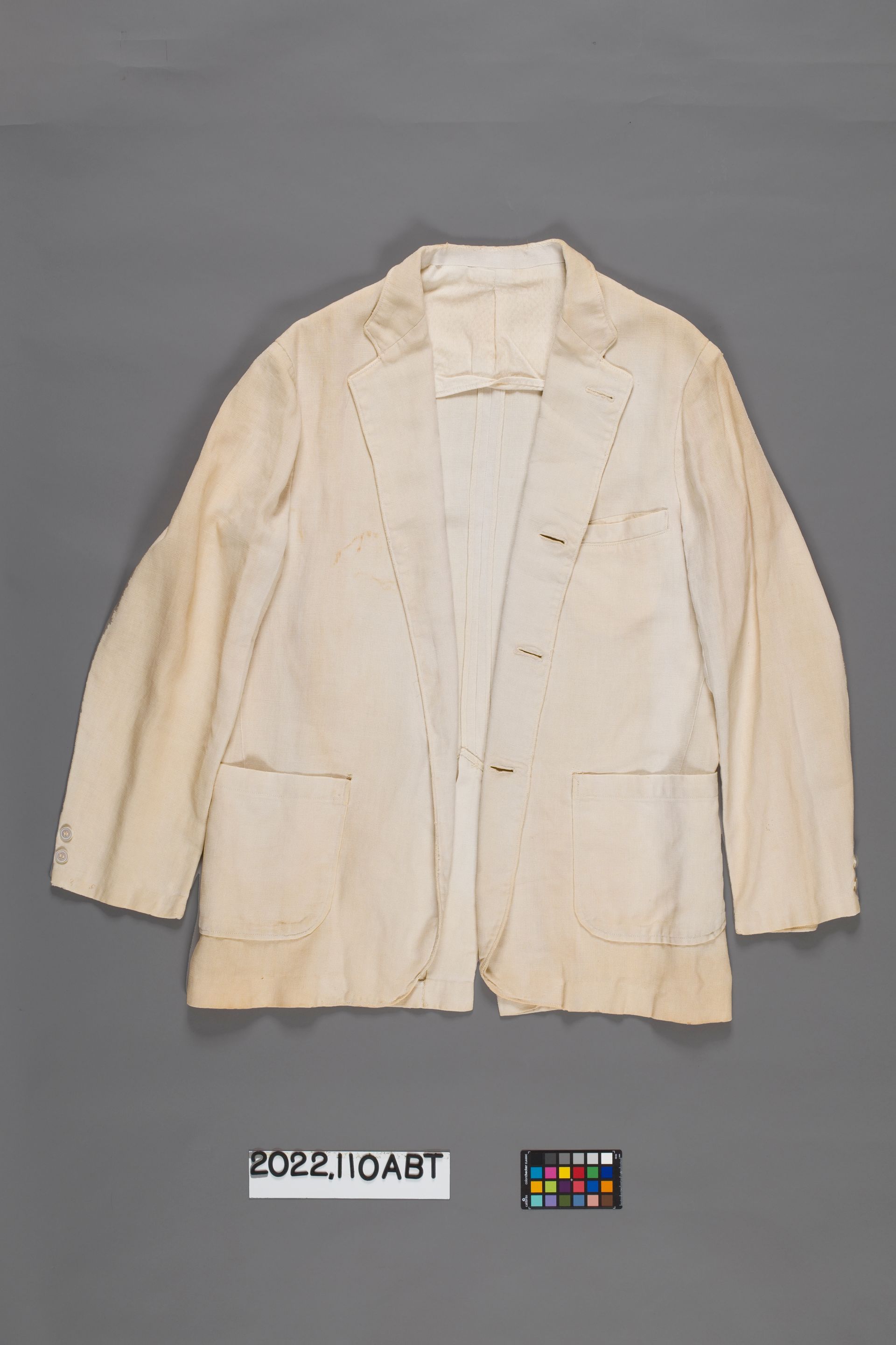 A light-colored, three-button blazer with two front pockets centered on a plain gray background.