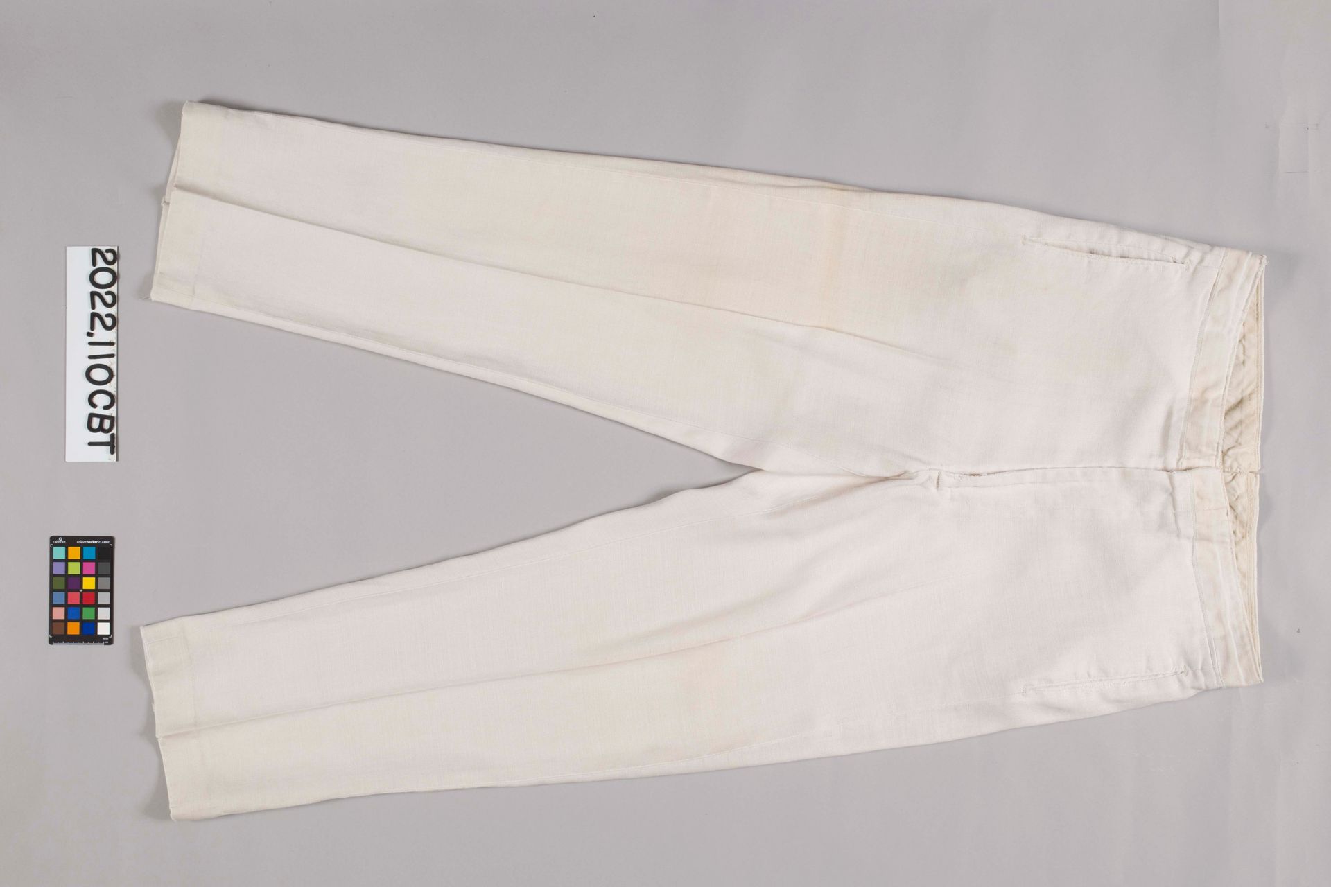 Off-white, straight-leg trousers laid flat on a neutral background, with a small color-reference card on the left.