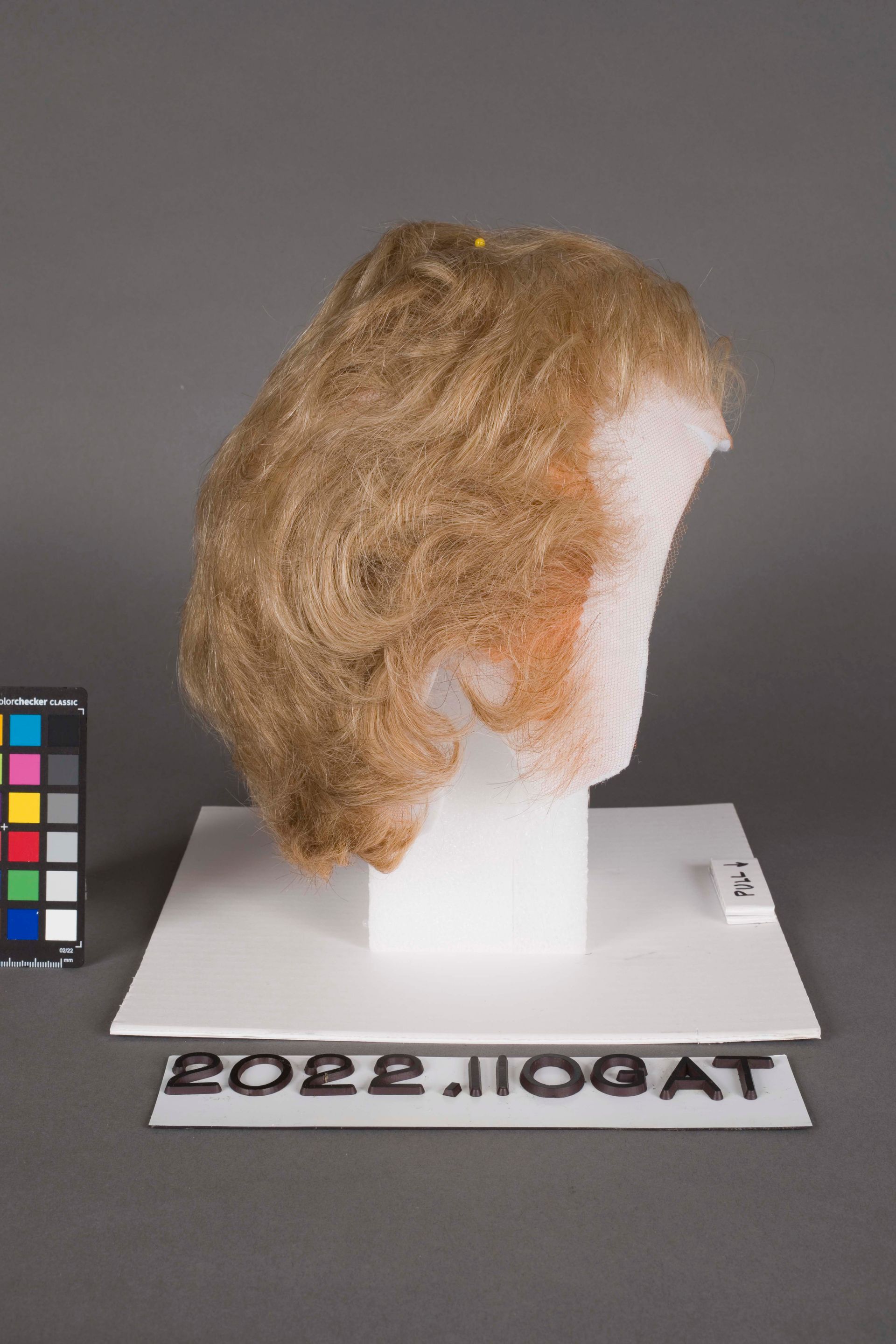 A light brown wig styled in loose waves displayed on a white foam head, next to a color calibration card.