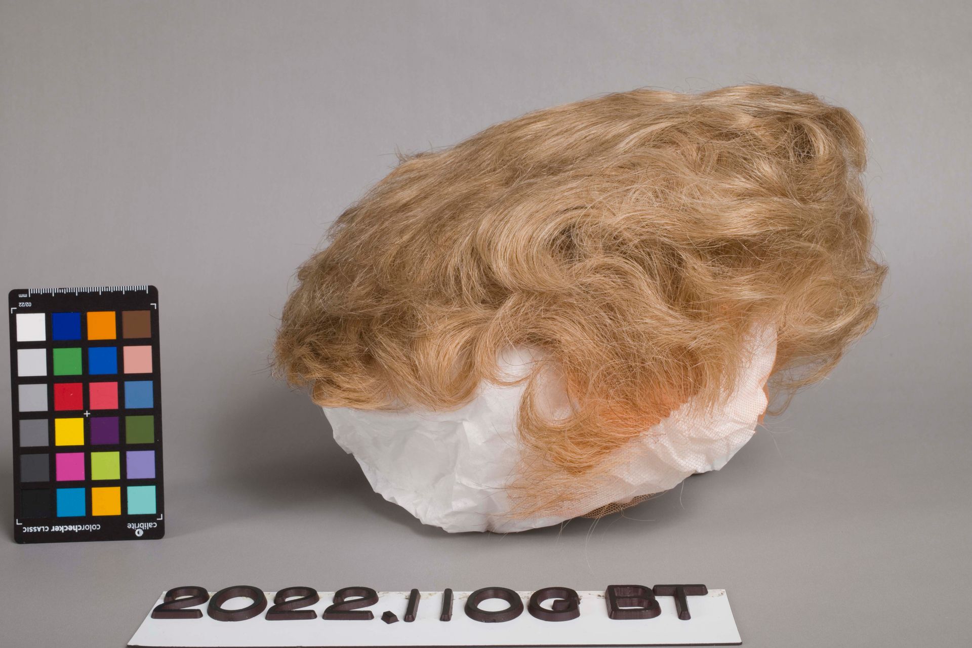 A light brown wig styled on a white foam head form next to a color calibration chart against a grey background.