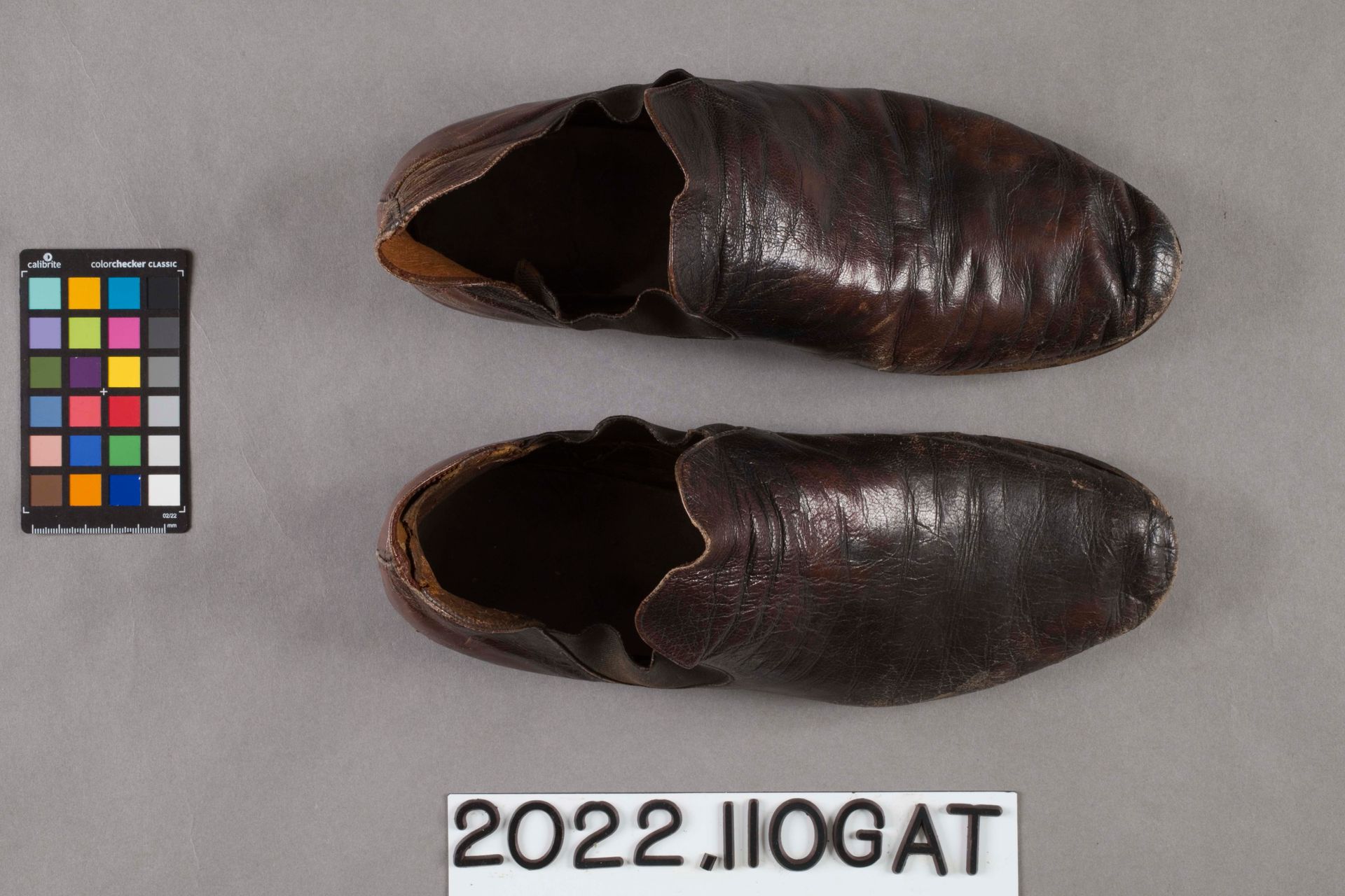 A pair of dark brown, vintage leather slip-on shoes viewed from above, alongside a color calibration chart and a label.