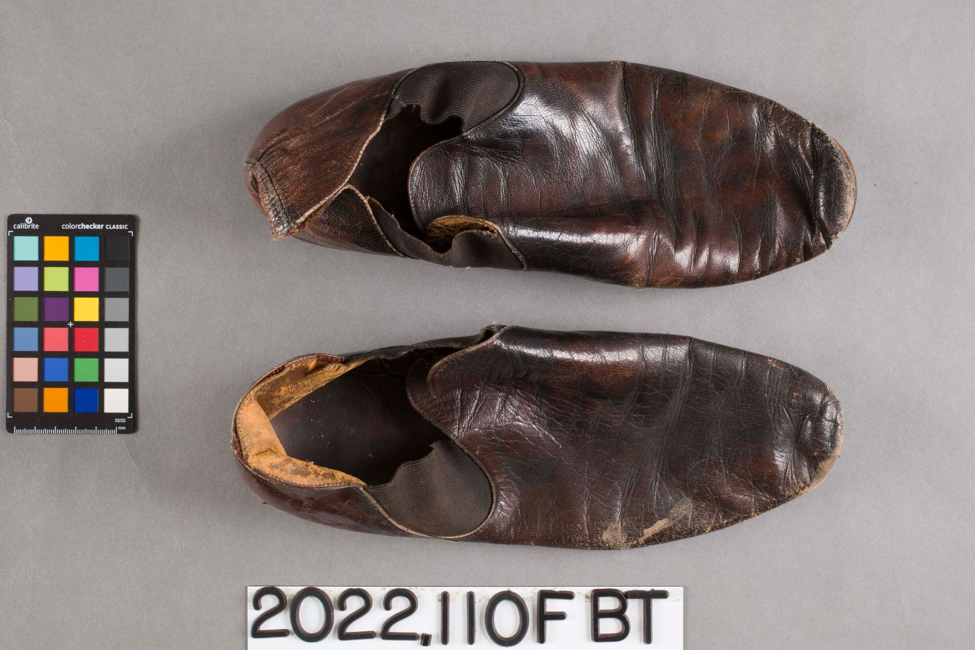 Two dark brown, worn leather slip-on shoes viewed from above, placed next to a color calibration card on a gray surface.