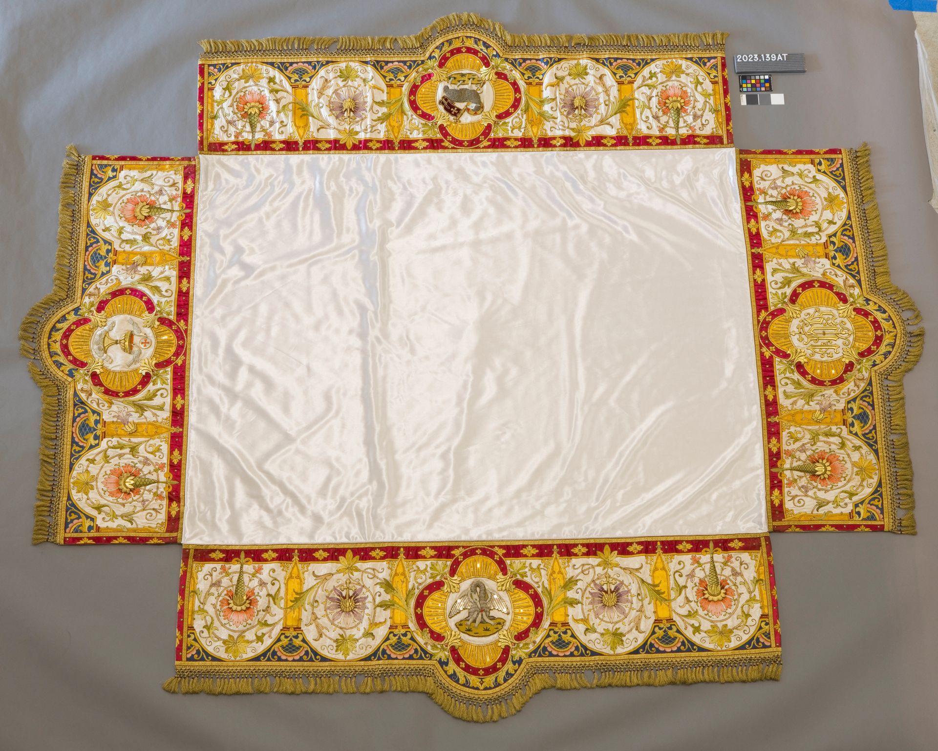 A white rectangular altar cloth bordered by gold and red panels featuring ornate floral embroidery and central crests.