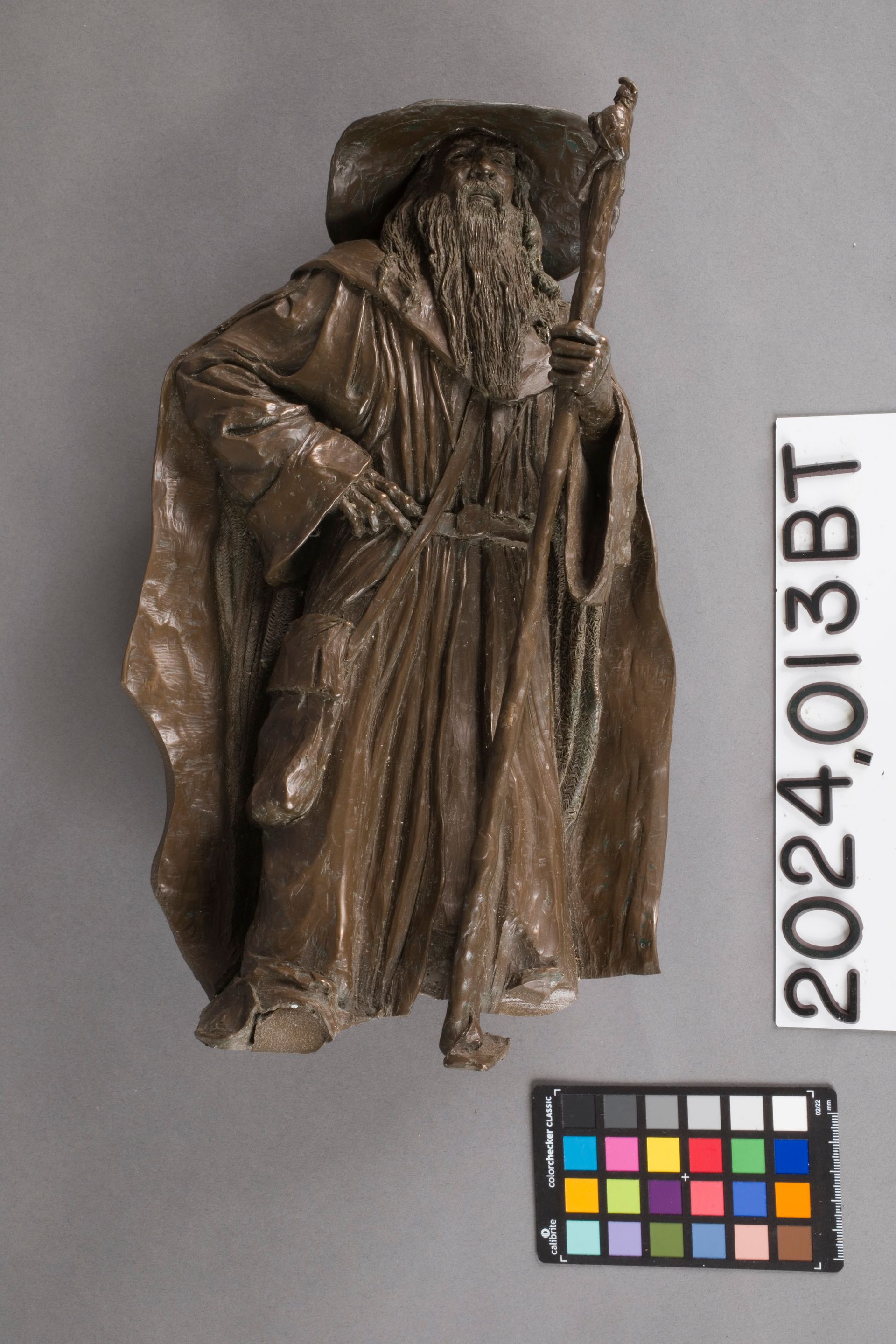 A brown, textured figurine of a cloaked, bearded figure in a wide-brimmed hat holding a tall wooden staff.