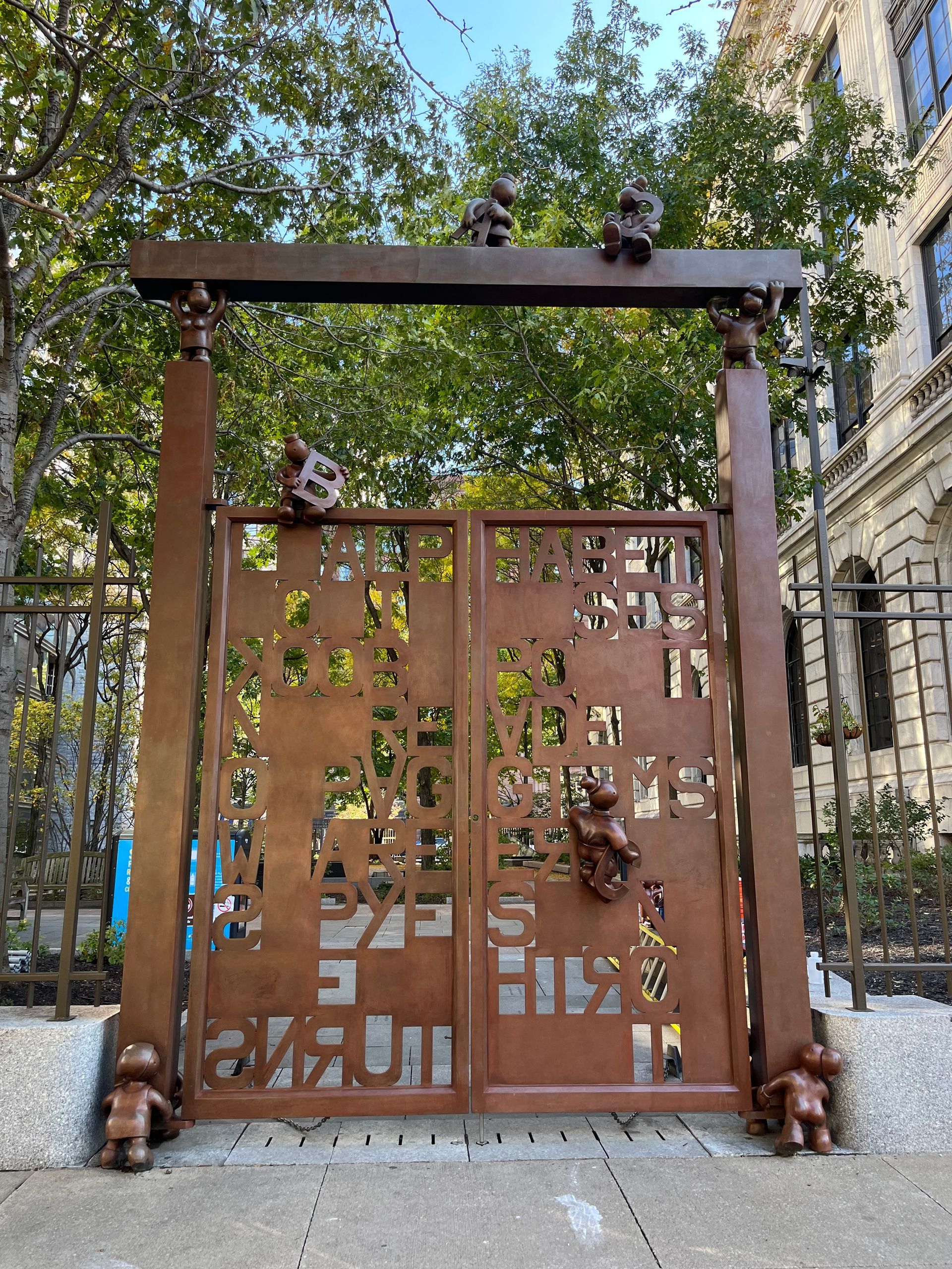 A weathered metal gate in an urban park, cut with abstract patterns and featuring small bronze figures climbing on it.