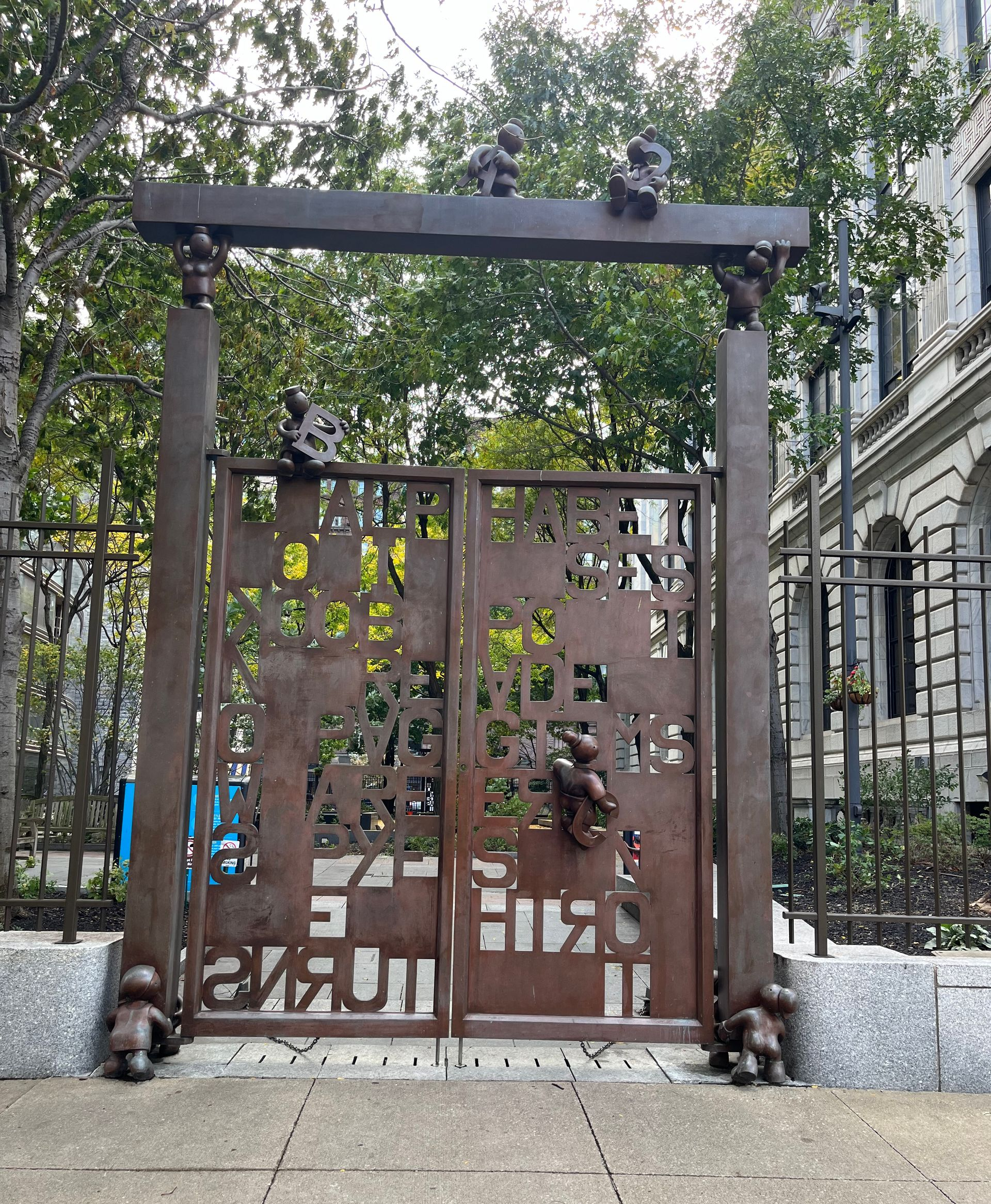 A brown metal gate featuring cut-out letters and small human-like figures climbing its frame and top in a park setting.
