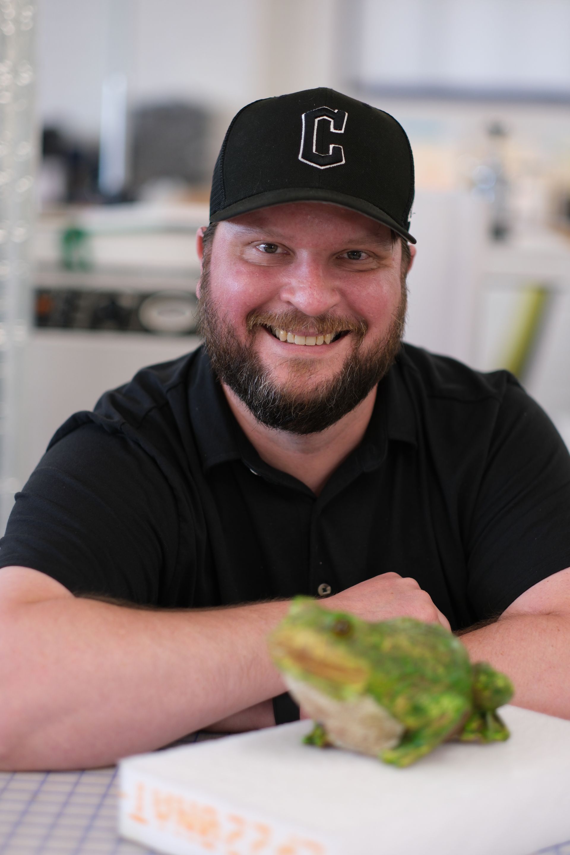 A man wearing a black hat is sitting next to a green frog.