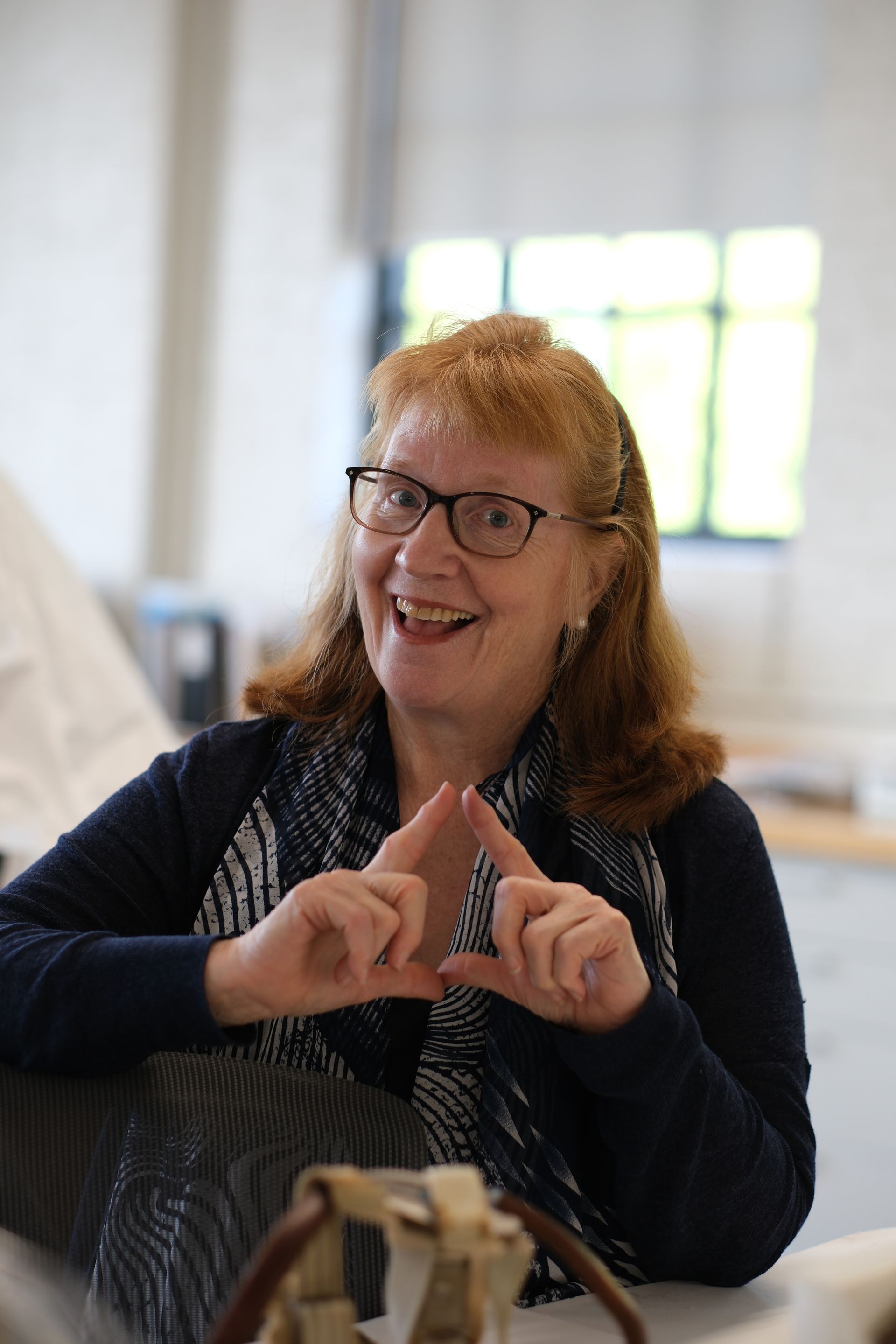 A woman wearing glasses and a scarf is sitting at a table making a heart with her hands.