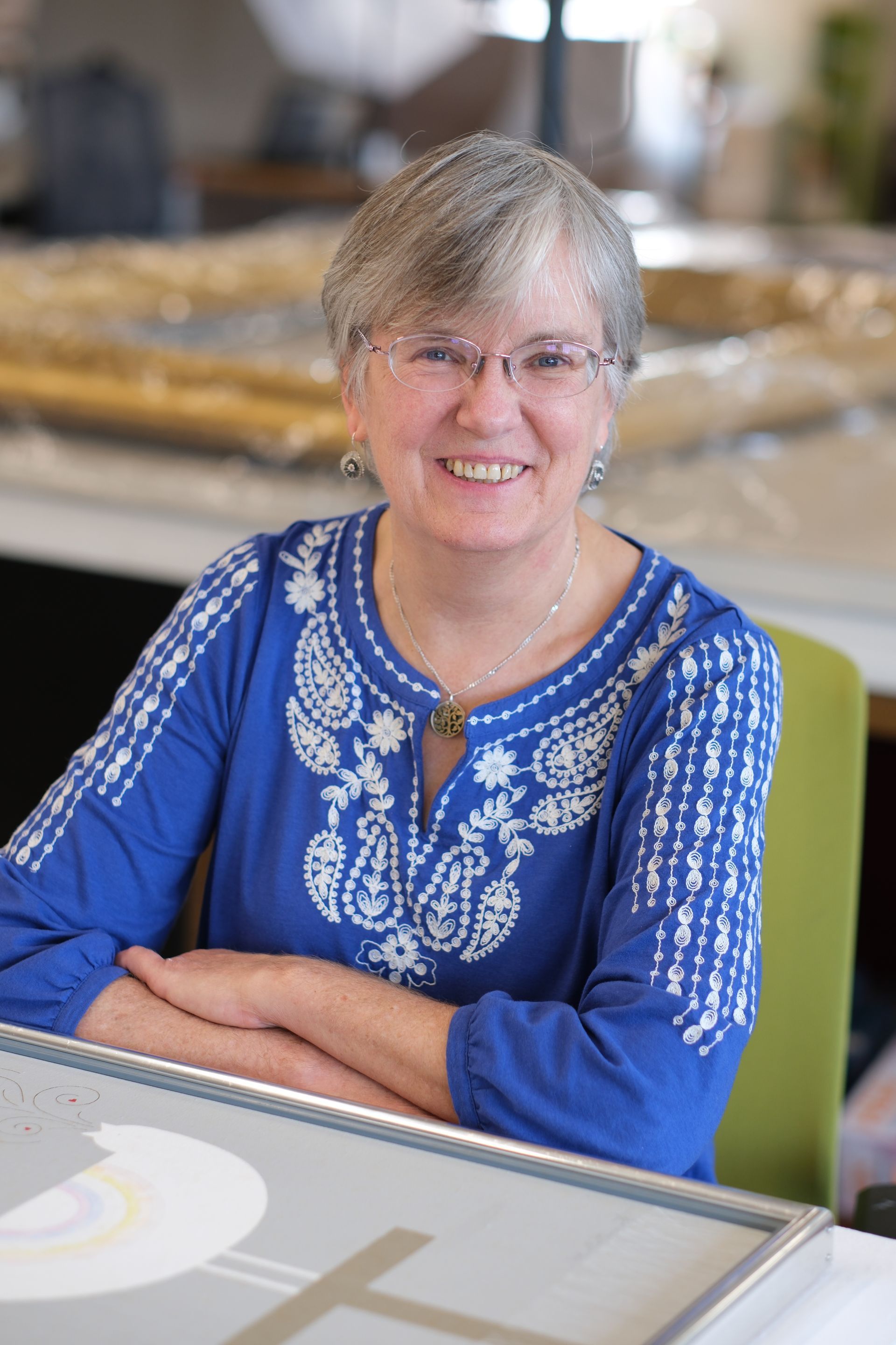 A woman in a blue shirt and glasses is sitting at a table with her arms crossed.