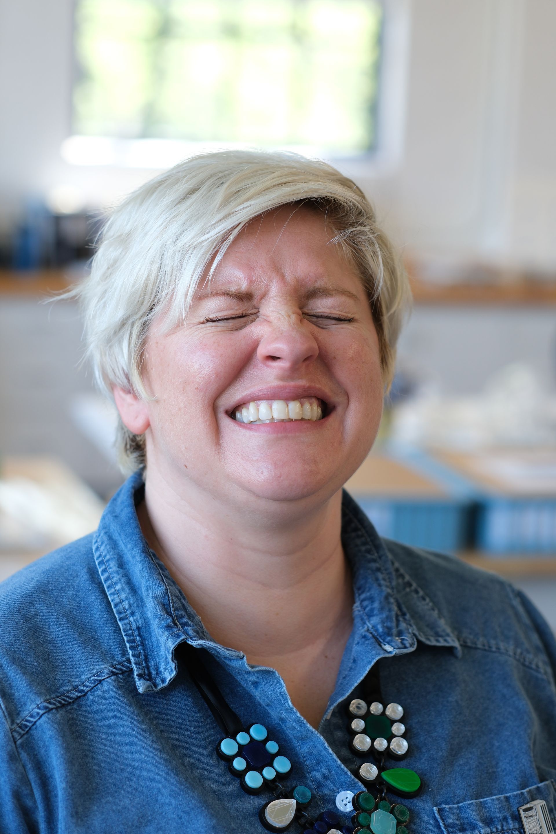 A woman is smiling with her eyes closed and wearing a denim shirt.