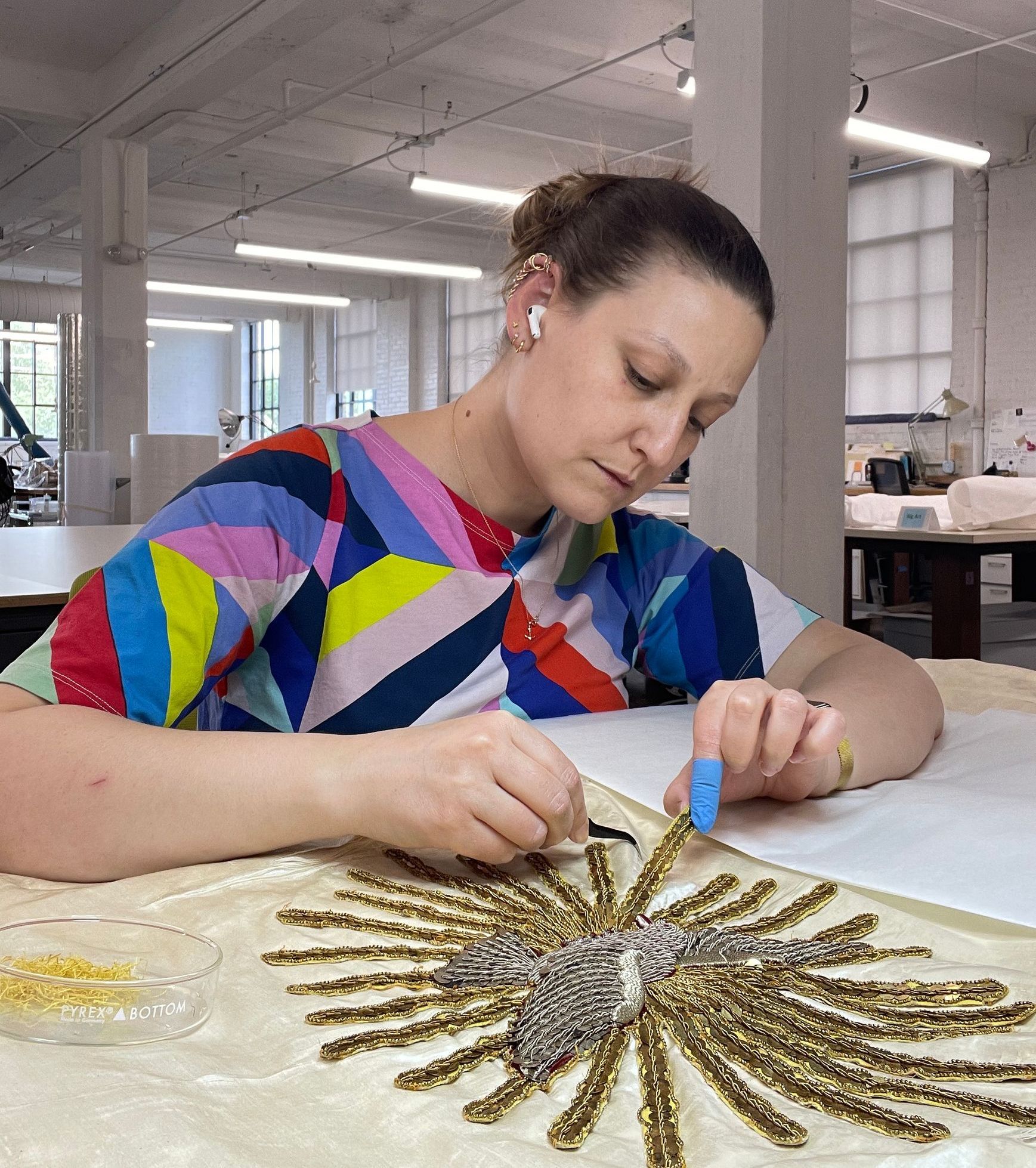 A woman is sitting at a table working on a piece of embroidery.