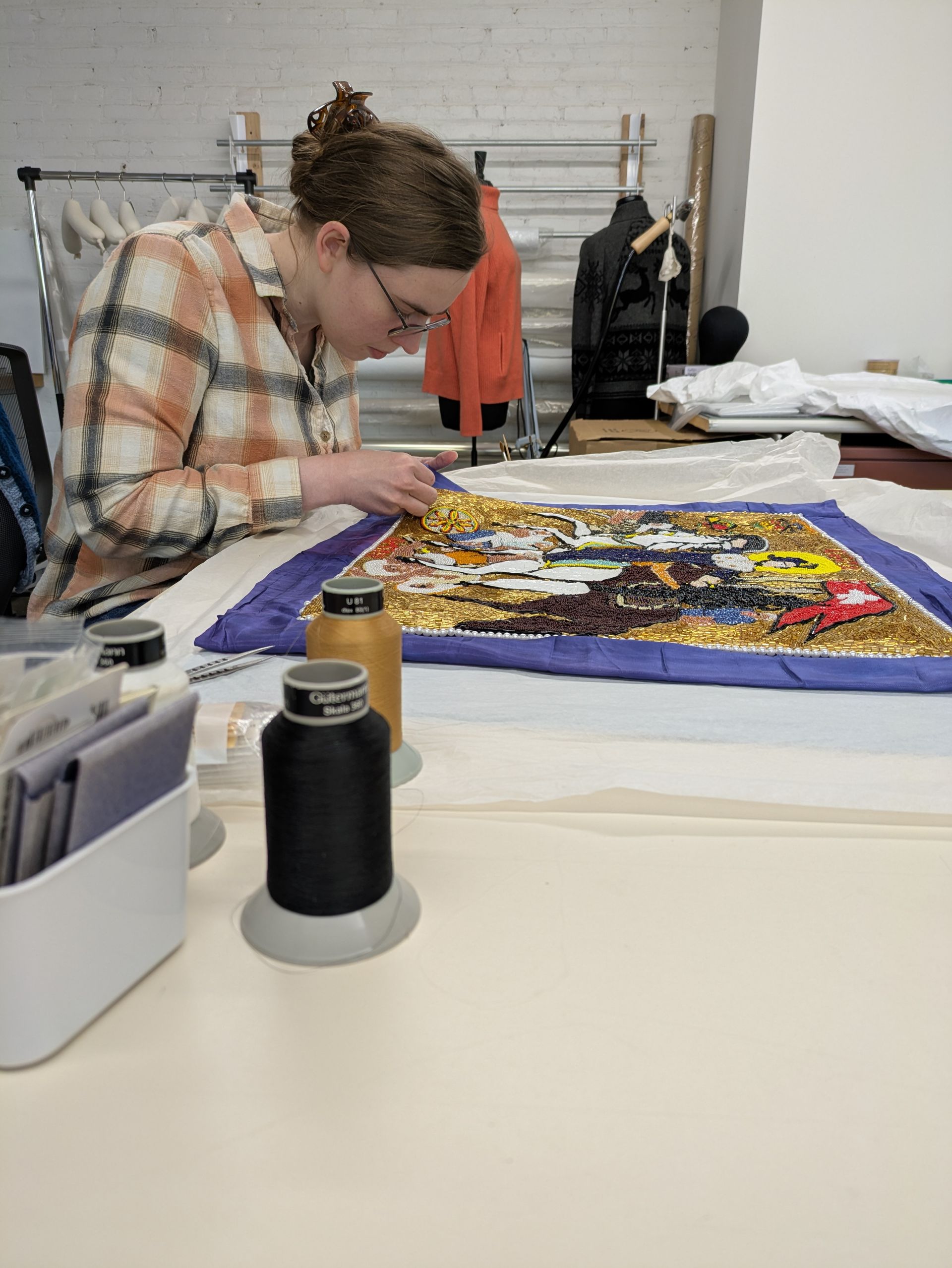 A woman is sitting at a table working on a piece of embroidery.