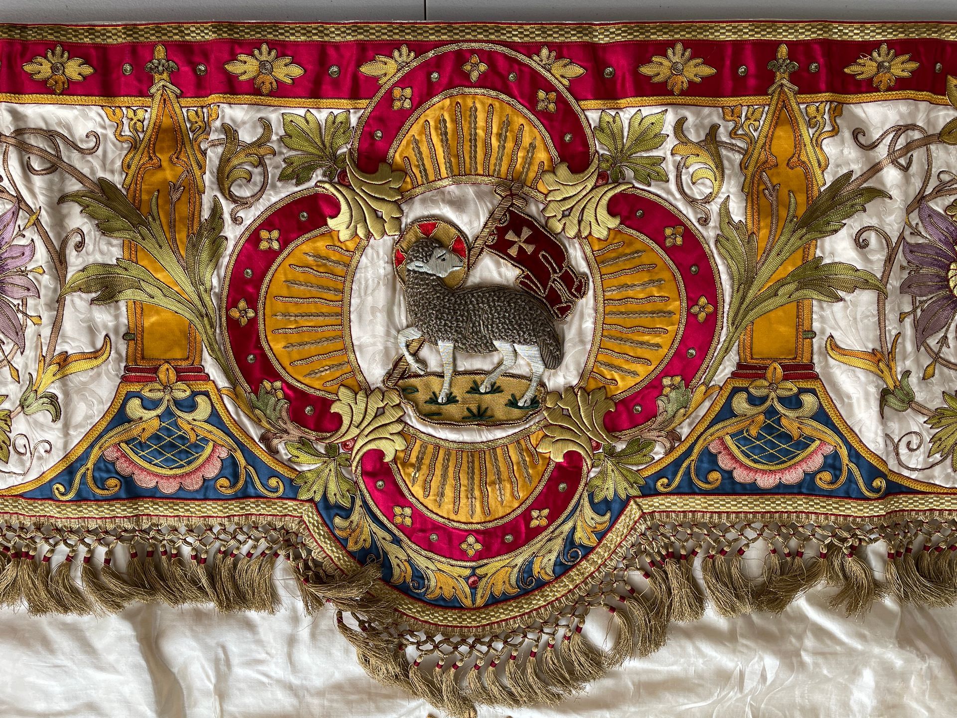 Ornate red, gold, and white textile featuring an embroidered Agnus Dei (Lamb of God) with a cross and fringe trim.