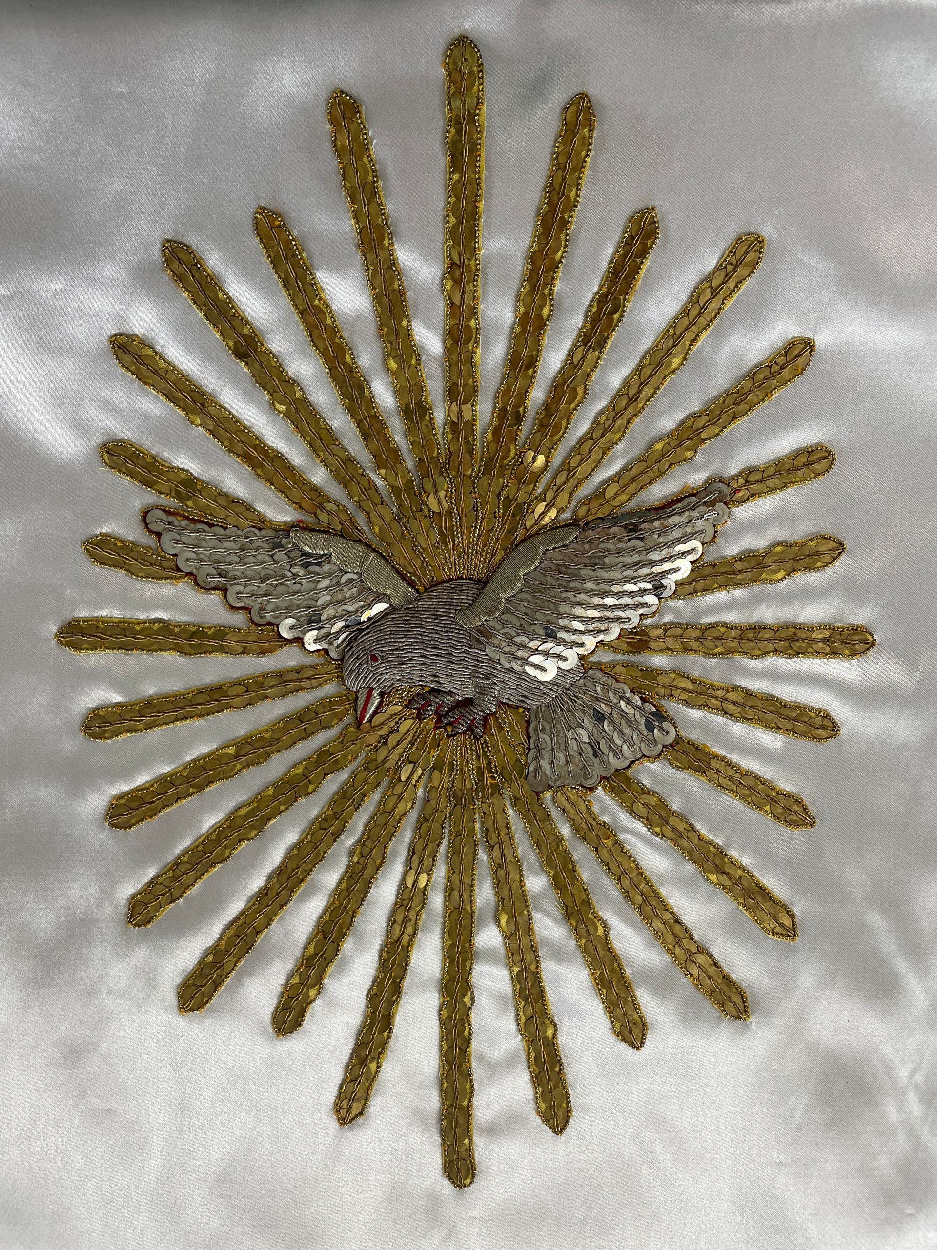 A silver dove centered within a radiant golden starburst, embroidered on a white satin background.