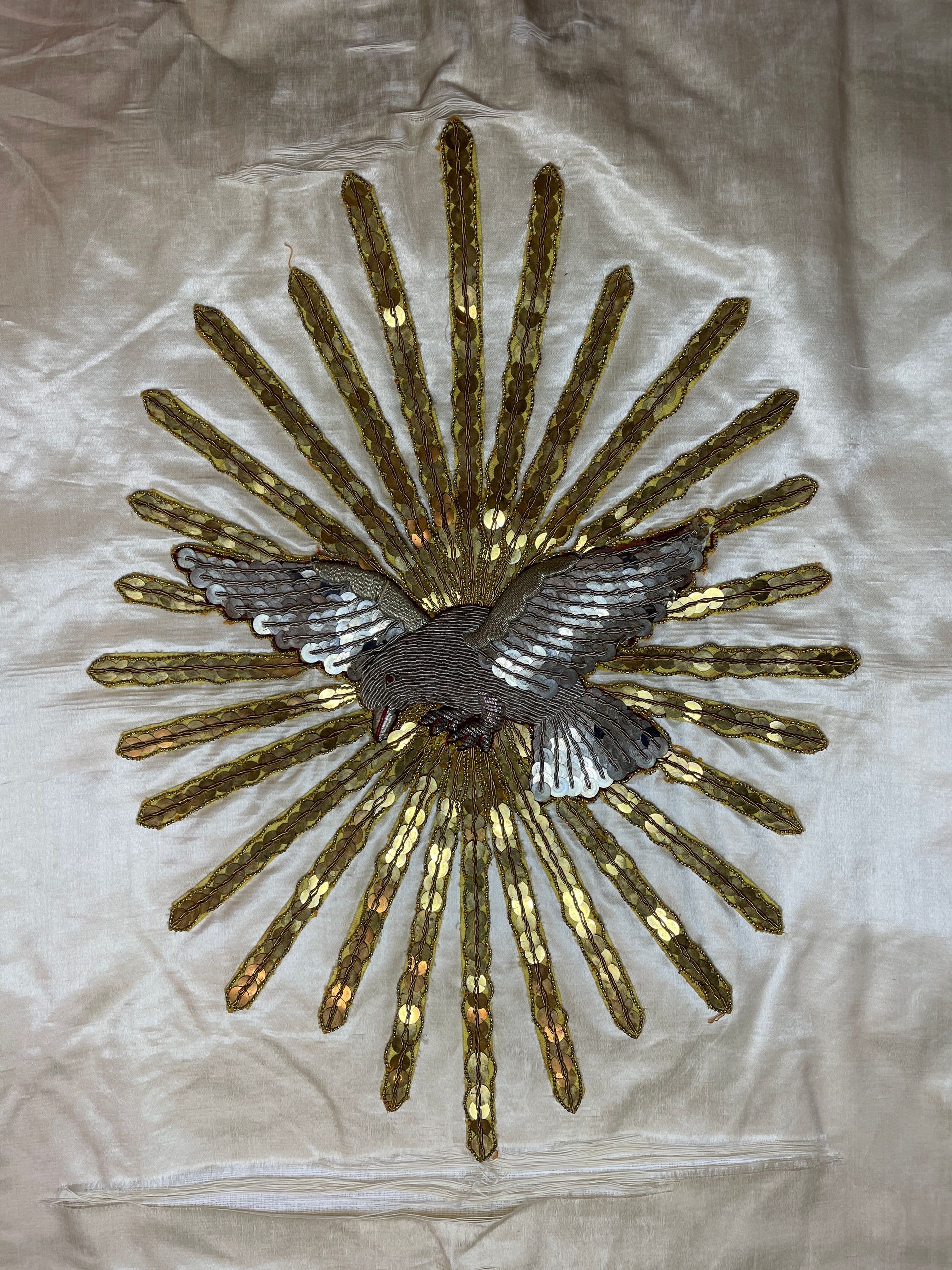A silver dove embroidered in thread and sequins, centered within a sunburst of gold sequin rays on a pale silk fabric.