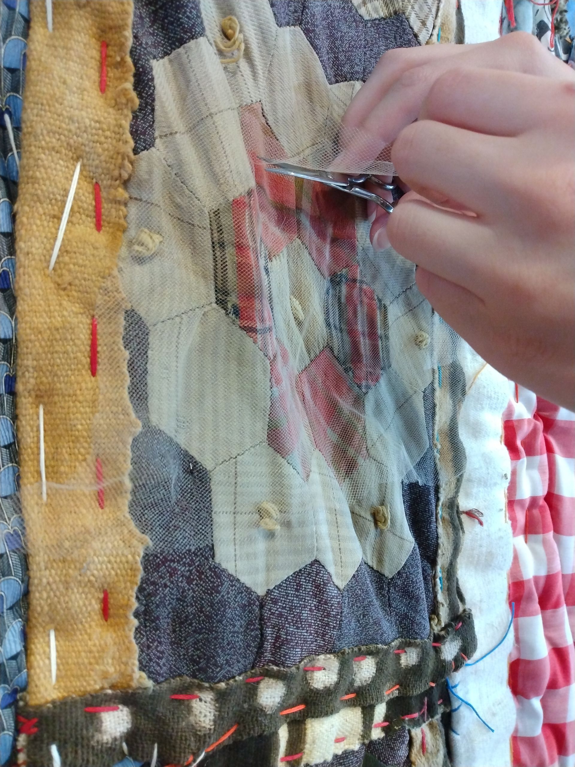 A hand uses small scissors to trim threads on a patchwork quilt featuring hexagon-shaped fabric pieces.
