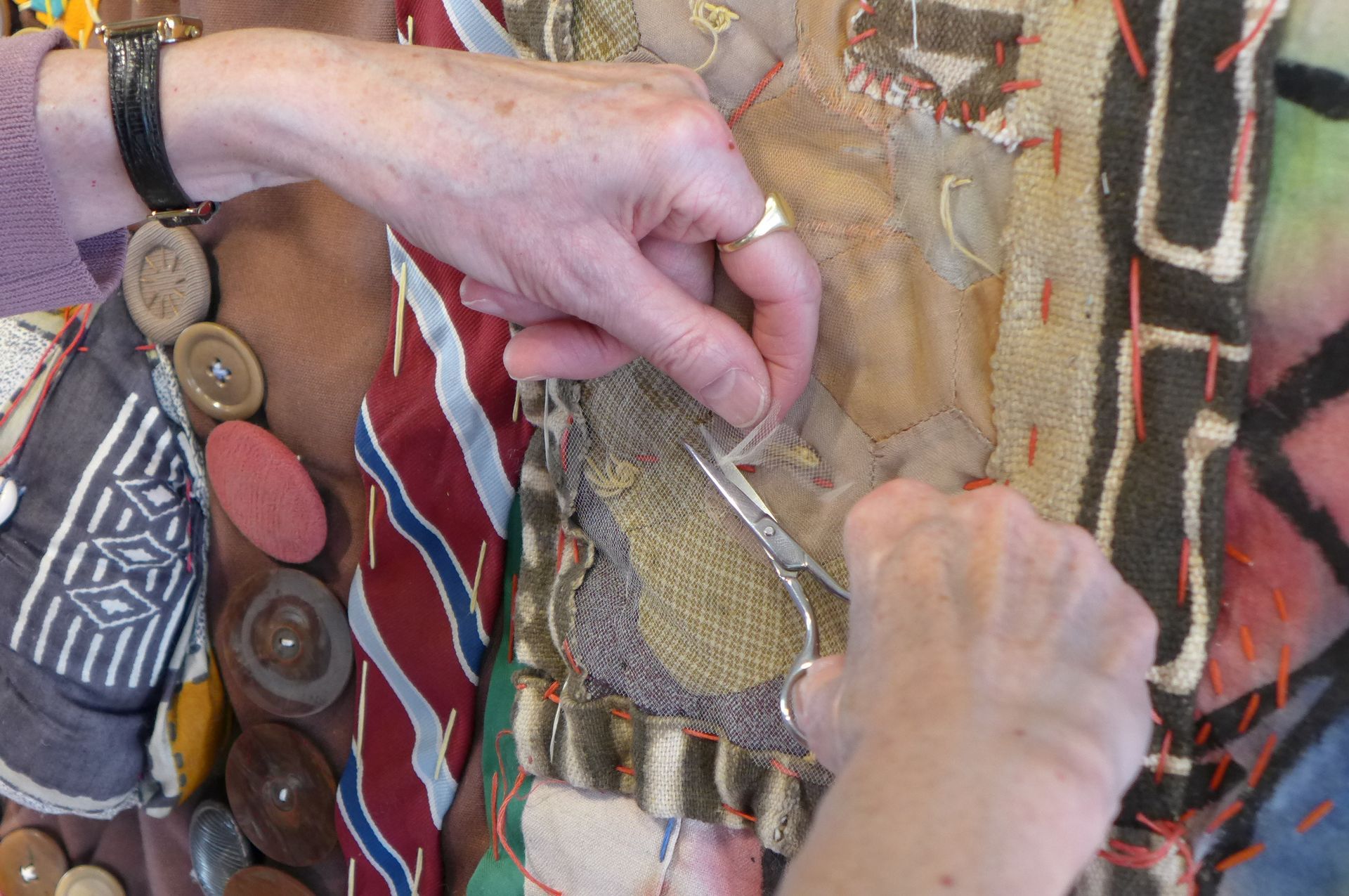 Hands work with scissors to trim a textured fabric quilt featuring buttons, various patterned cloths, and orange stitching.