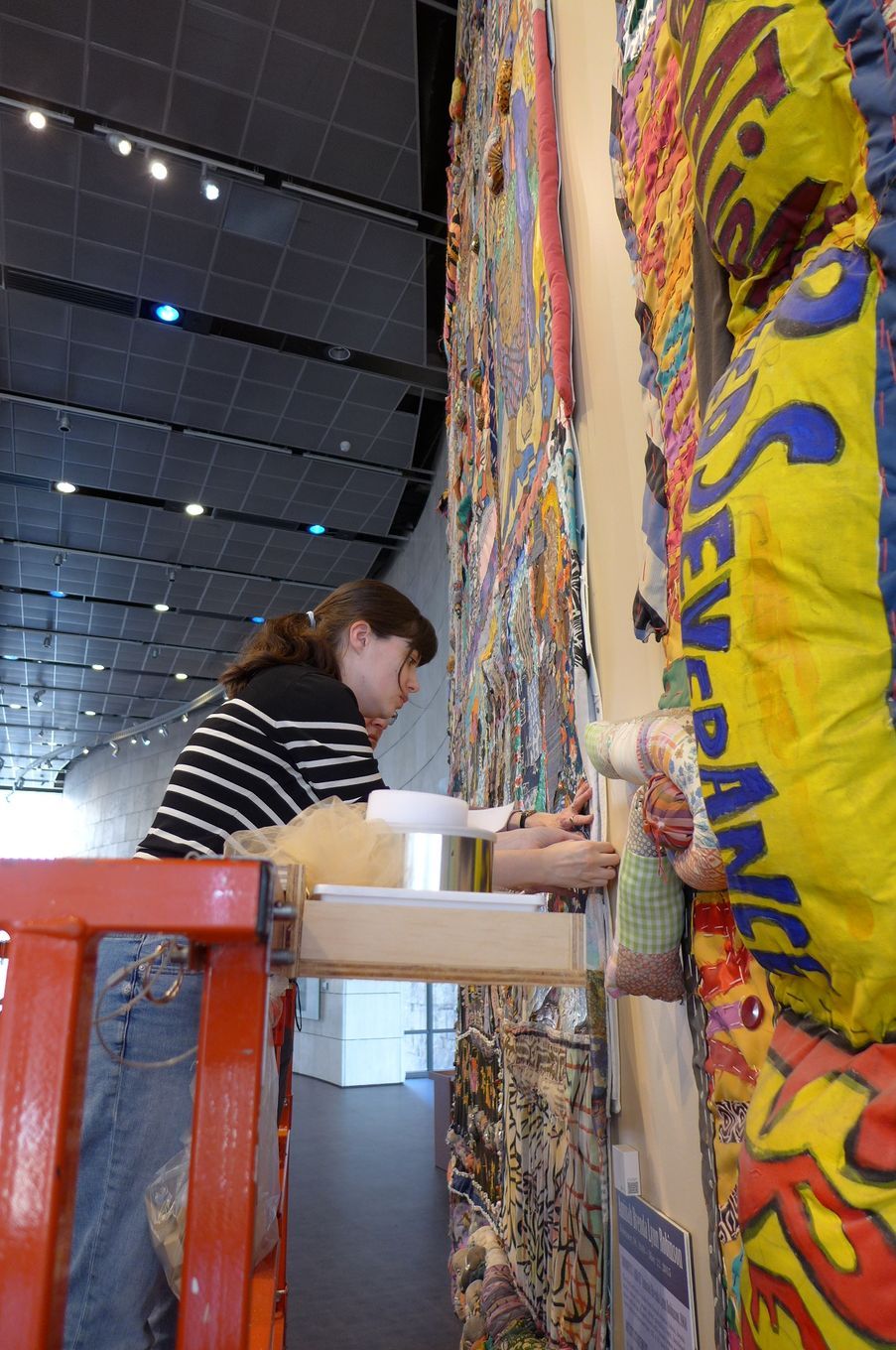 A person in a striped shirt stands on a red ladder, working to install a large, colorful, fabric-based art piece on a wall.
