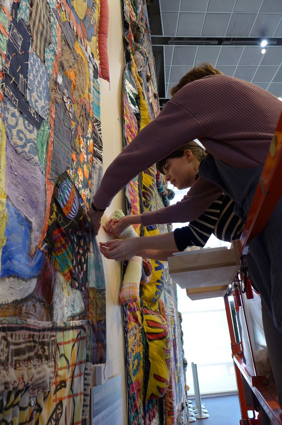 Two people on a ladder reach up to pin a large, colorful, patterned textile hanging on a gallery wall.