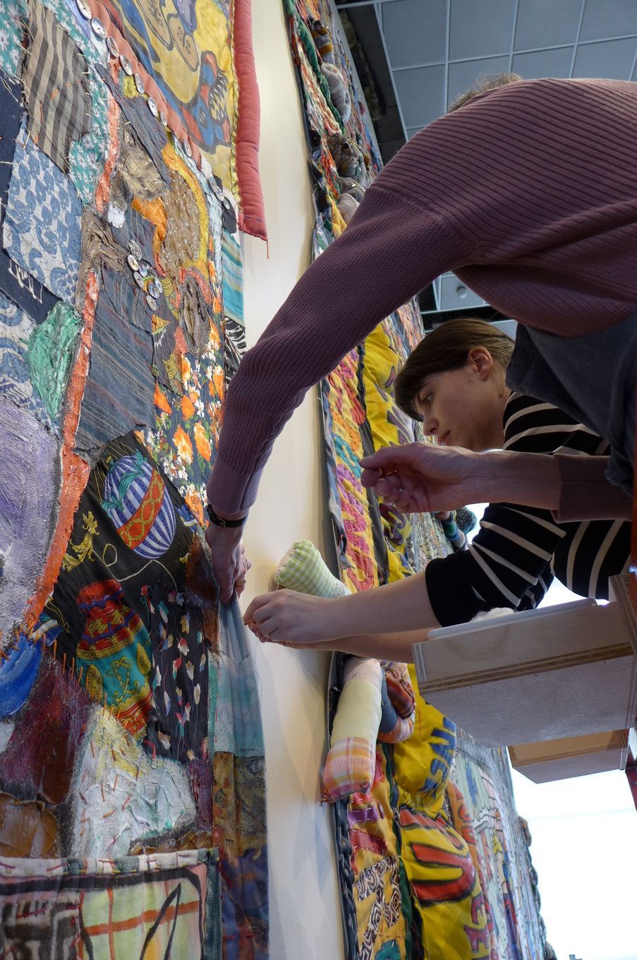 Two people work together to attach a large, colorful, patterned fabric tapestry to a plain white wall.