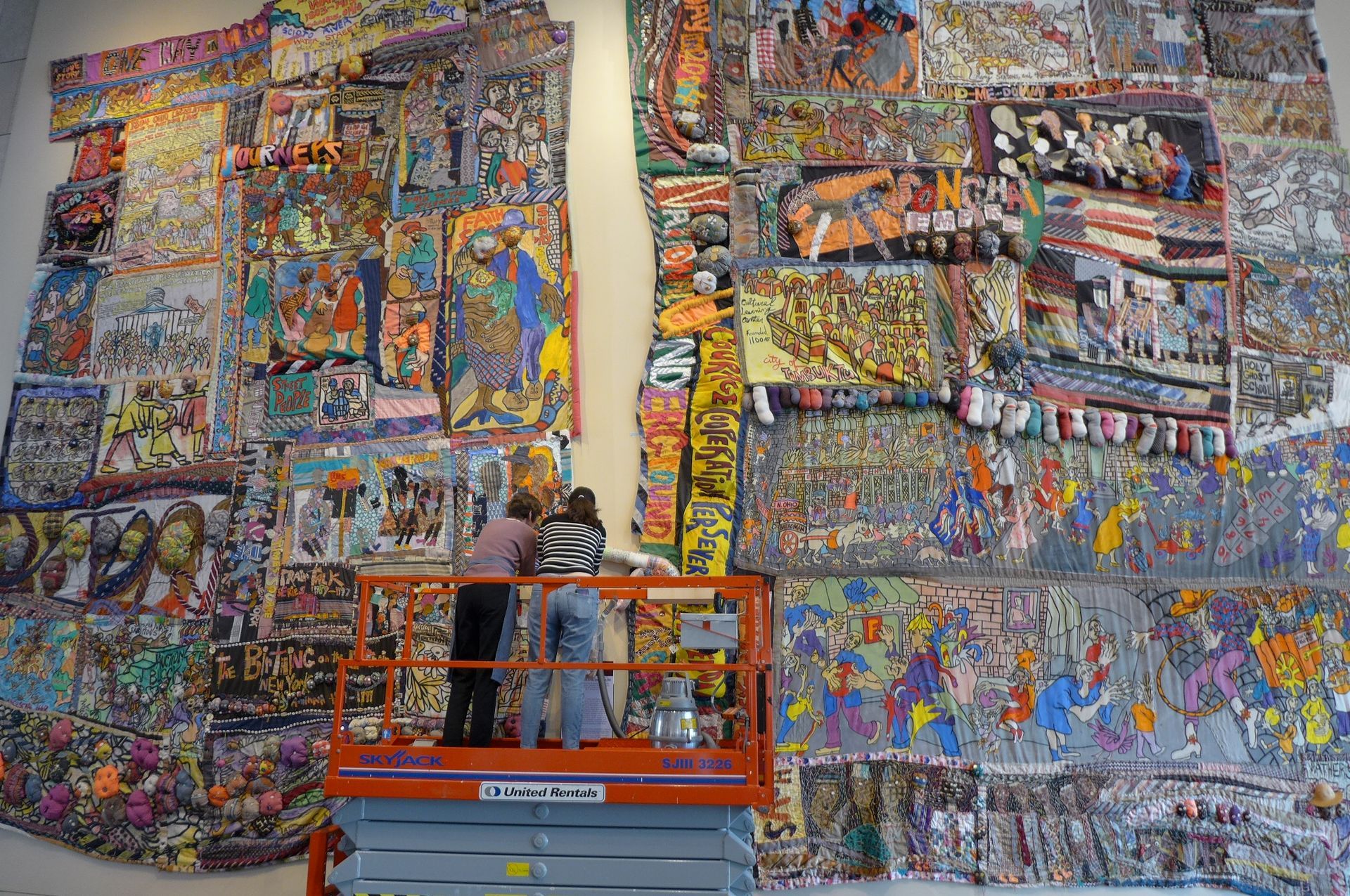 Two people on an orange scissor lift work on a large, colorful, multi-paneled mural covering a white gallery wall.