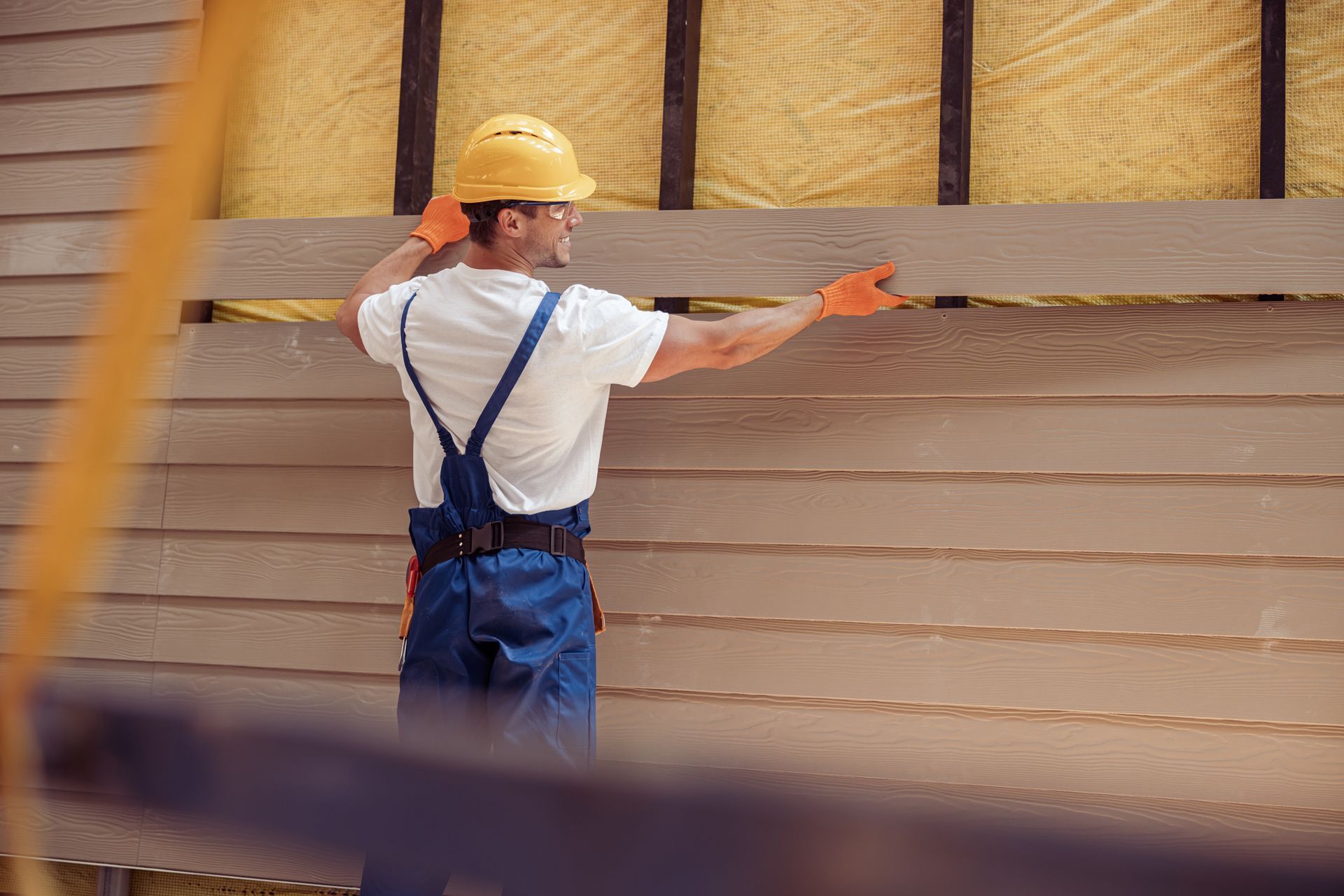 A construction worker in a hard hat and blue overalls installs horizontal siding on the exterior wall of a building.