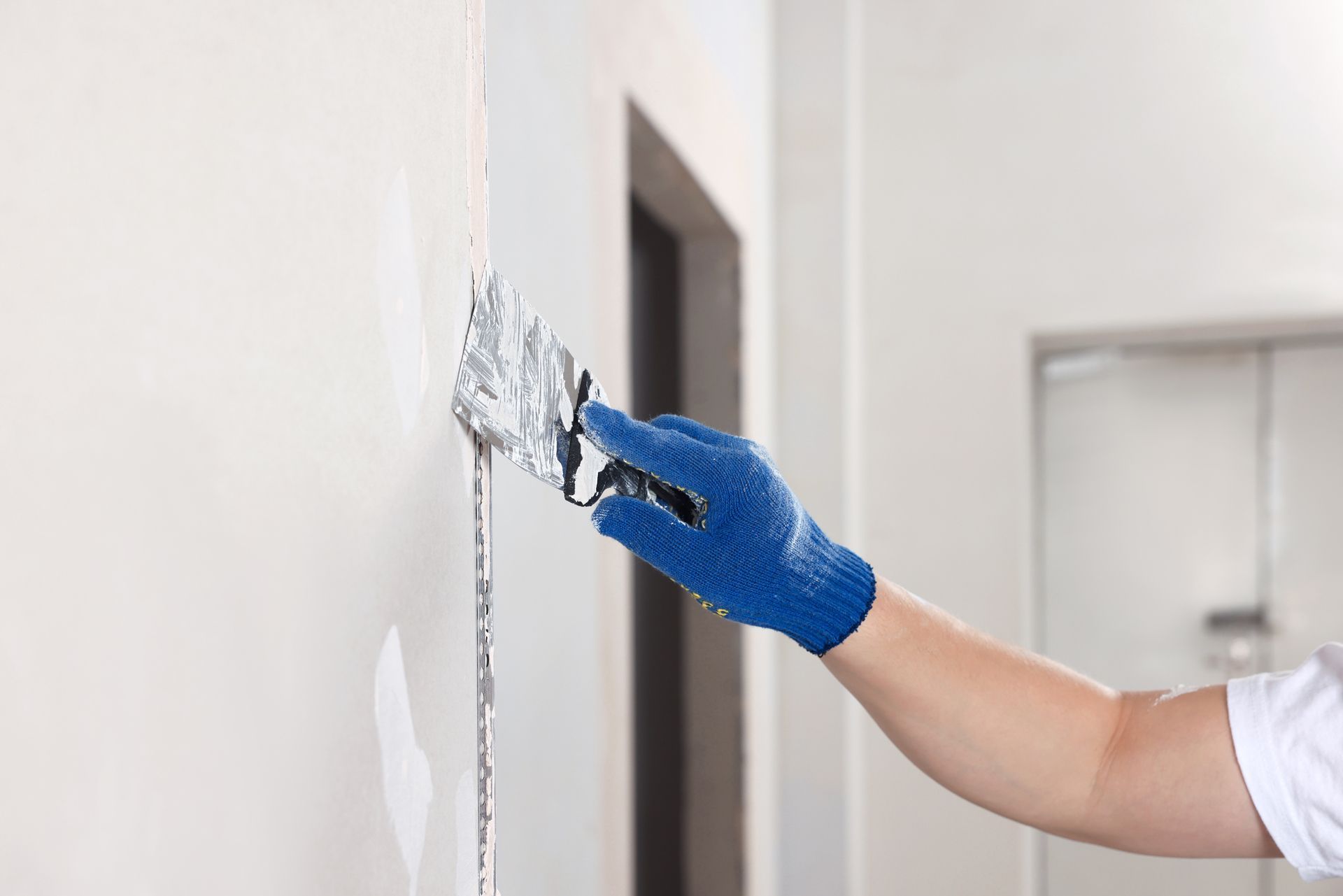 A person wearing blue gloves uses a metal putty knife to apply joint compound to a seam on a white wall.