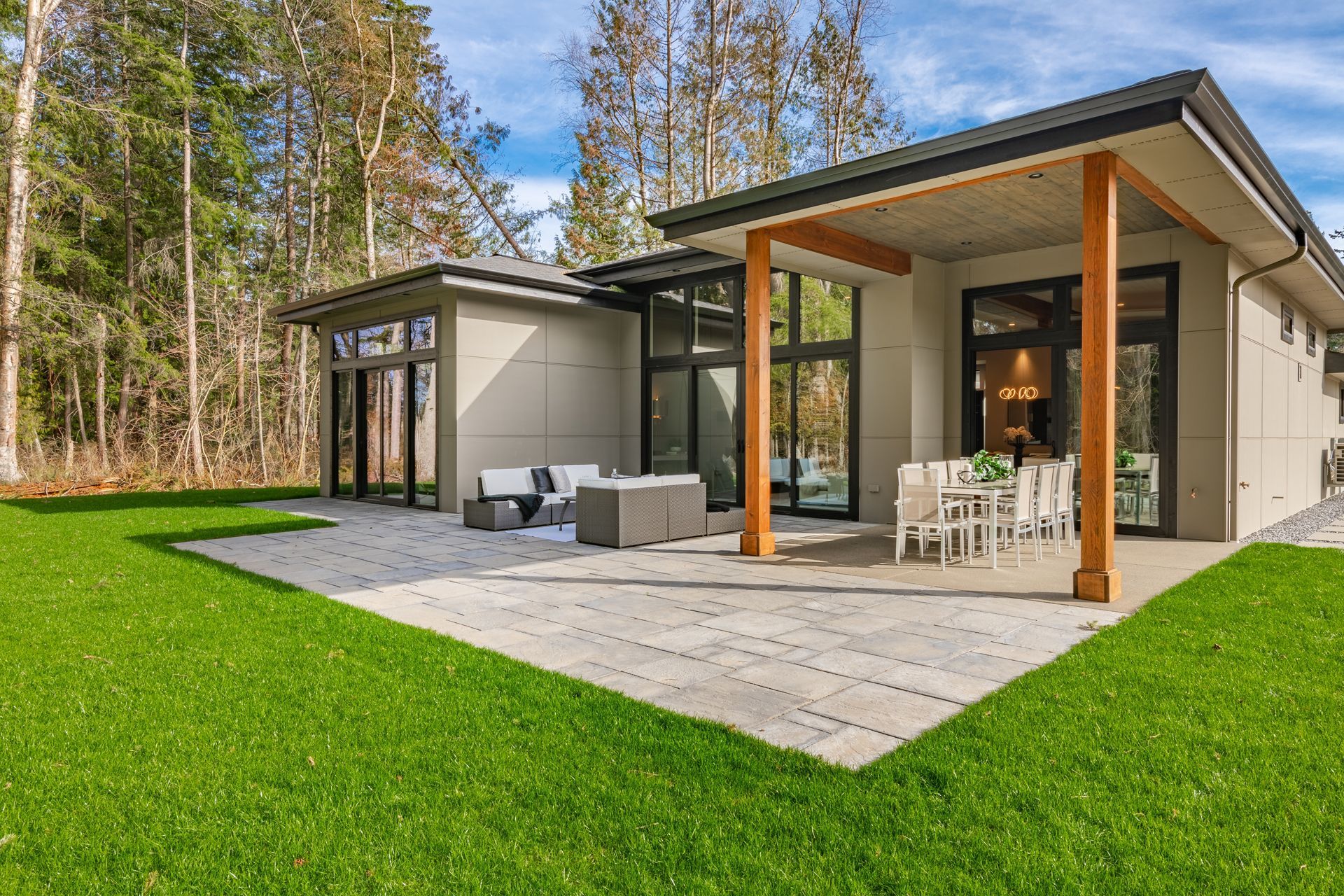 Modern house with a stone patio, outdoor seating, and large windows, surrounded by a forest and a grassy lawn.