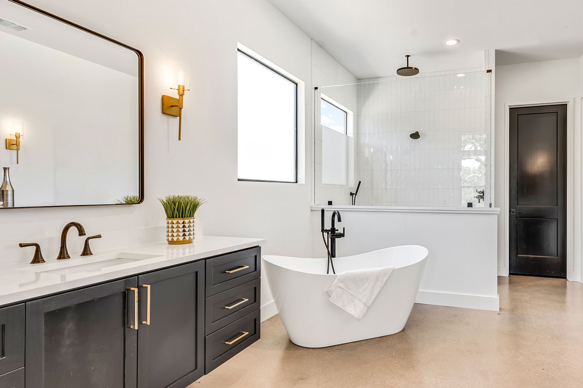 A modern, bright bathroom with dark charcoal cabinets, a white freestanding tub, and a glass-enclosed shower.