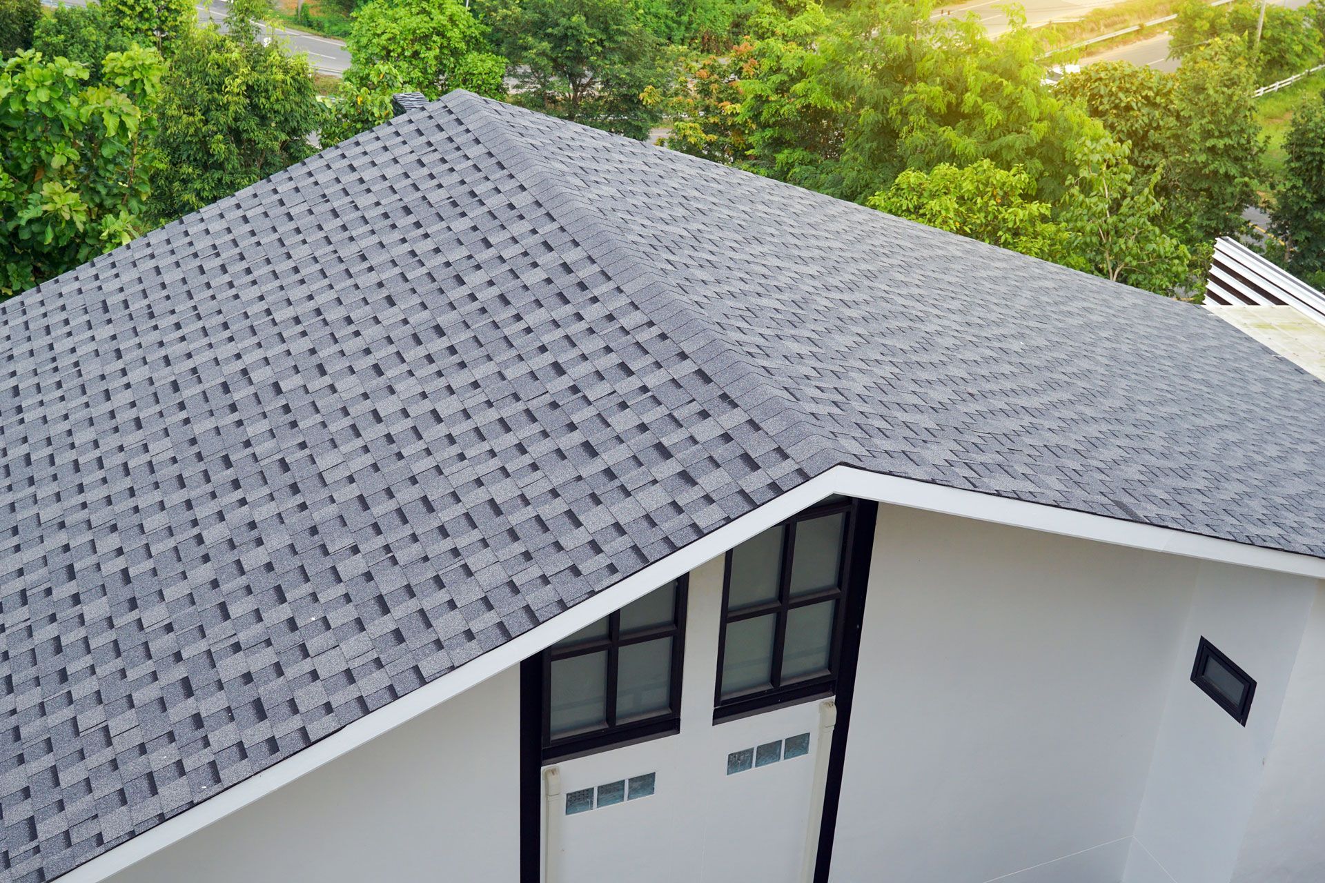 Gray shingled roof on a white house, viewed from above, surrounded by green trees.