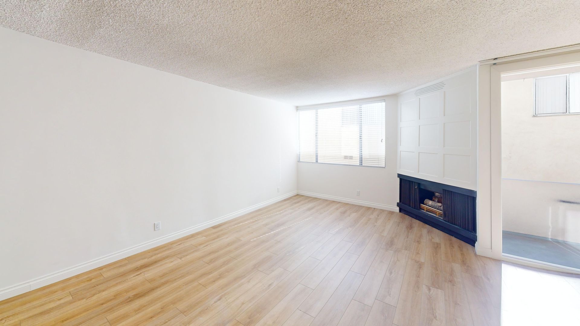 Empty living room with light wood floors, white walls, a window, and a dark fireplace.