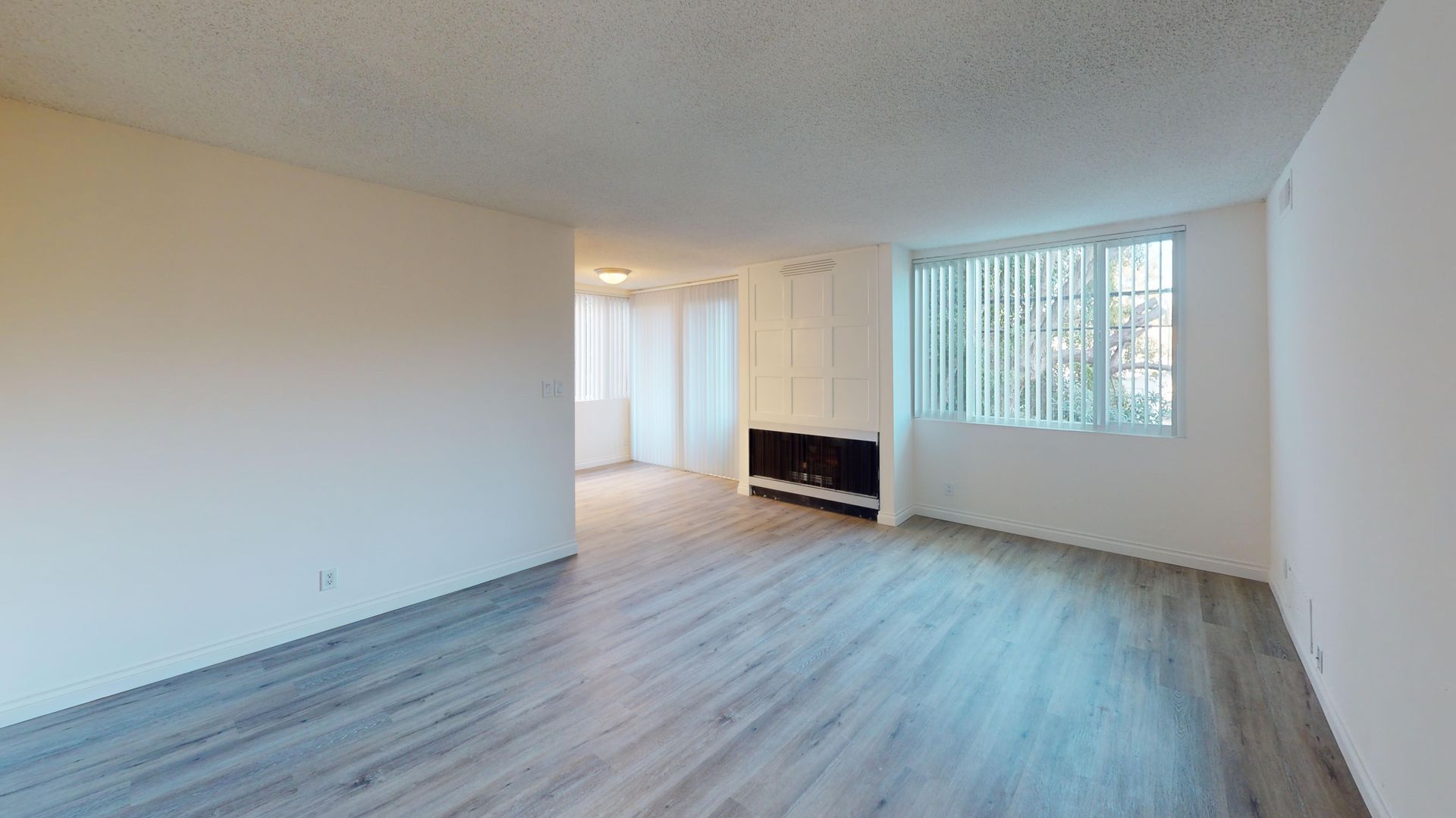 Empty living room with wood-look flooring, white walls, large window with blinds, and a built-in heater.