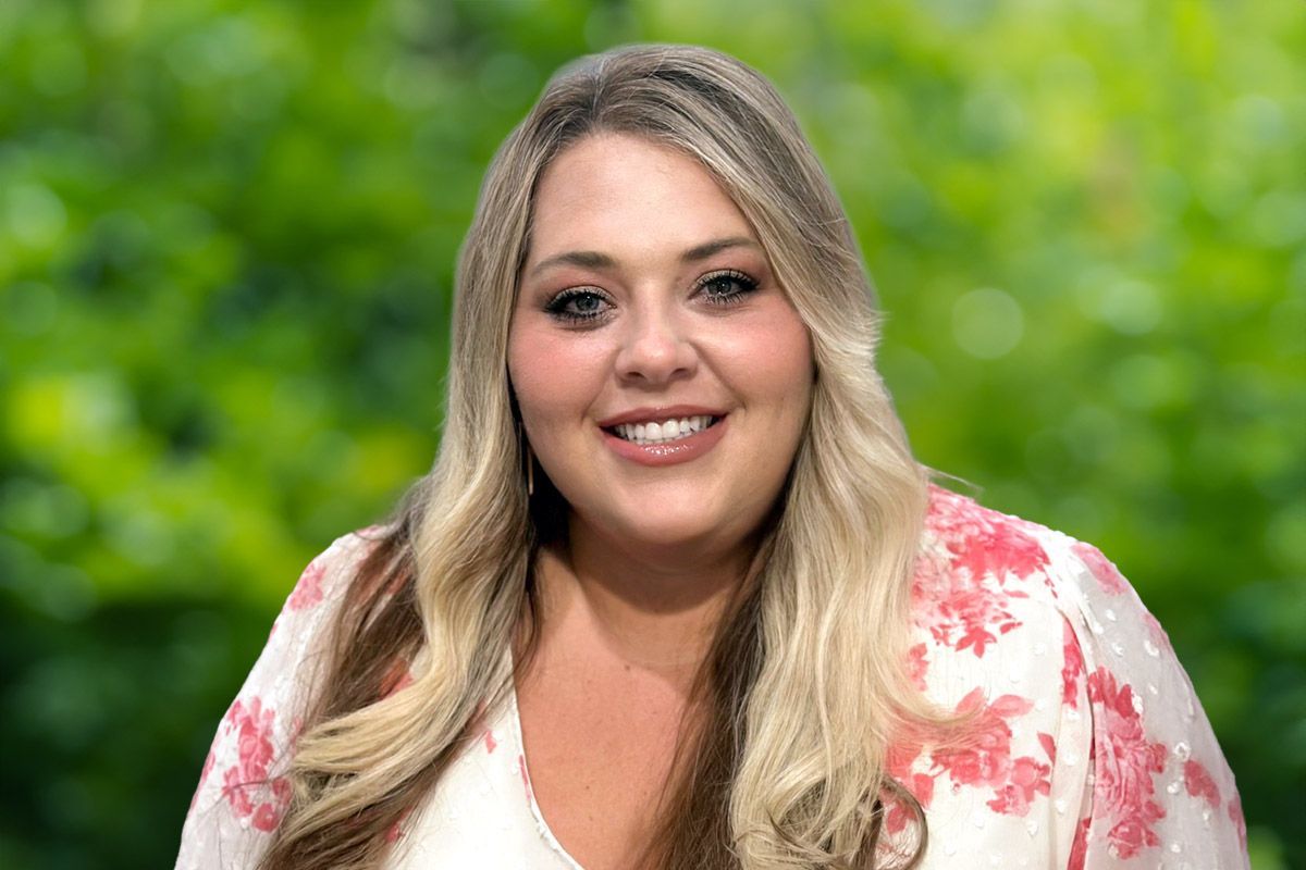 Woman with long blonde hair smiles outdoors, wearing a white and pink floral shirt against a green, blurred background.